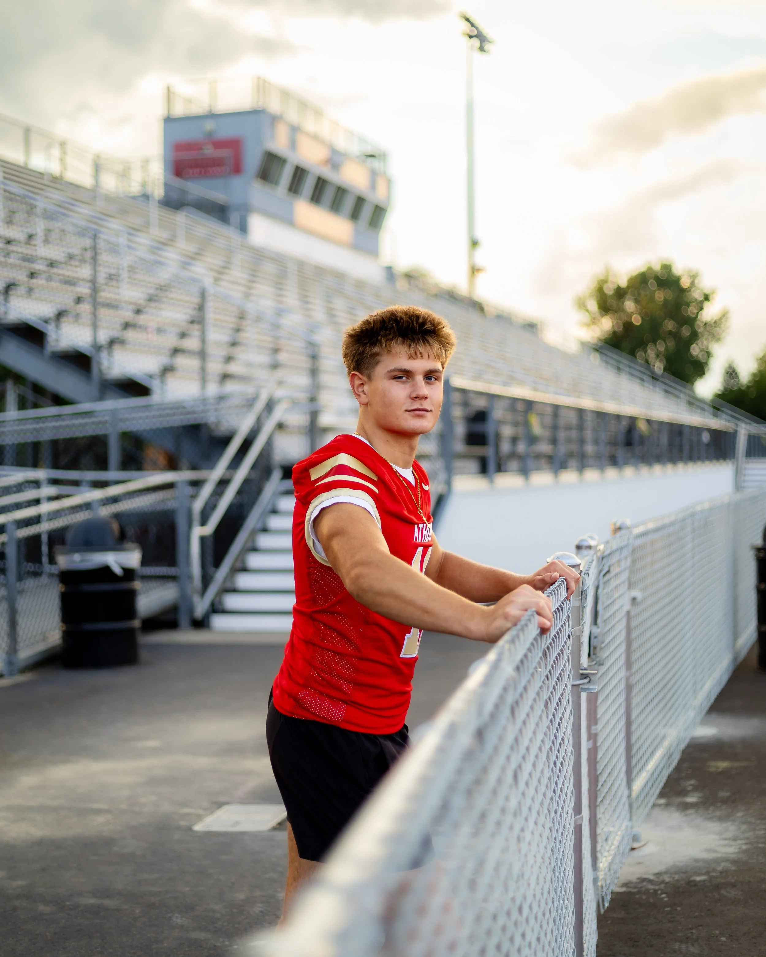 A young male athlete wearing a red sports jersey and black shorts standing behind a chain-link fence at a sports stadium, with bleachers and a control tower in the background during sunset.