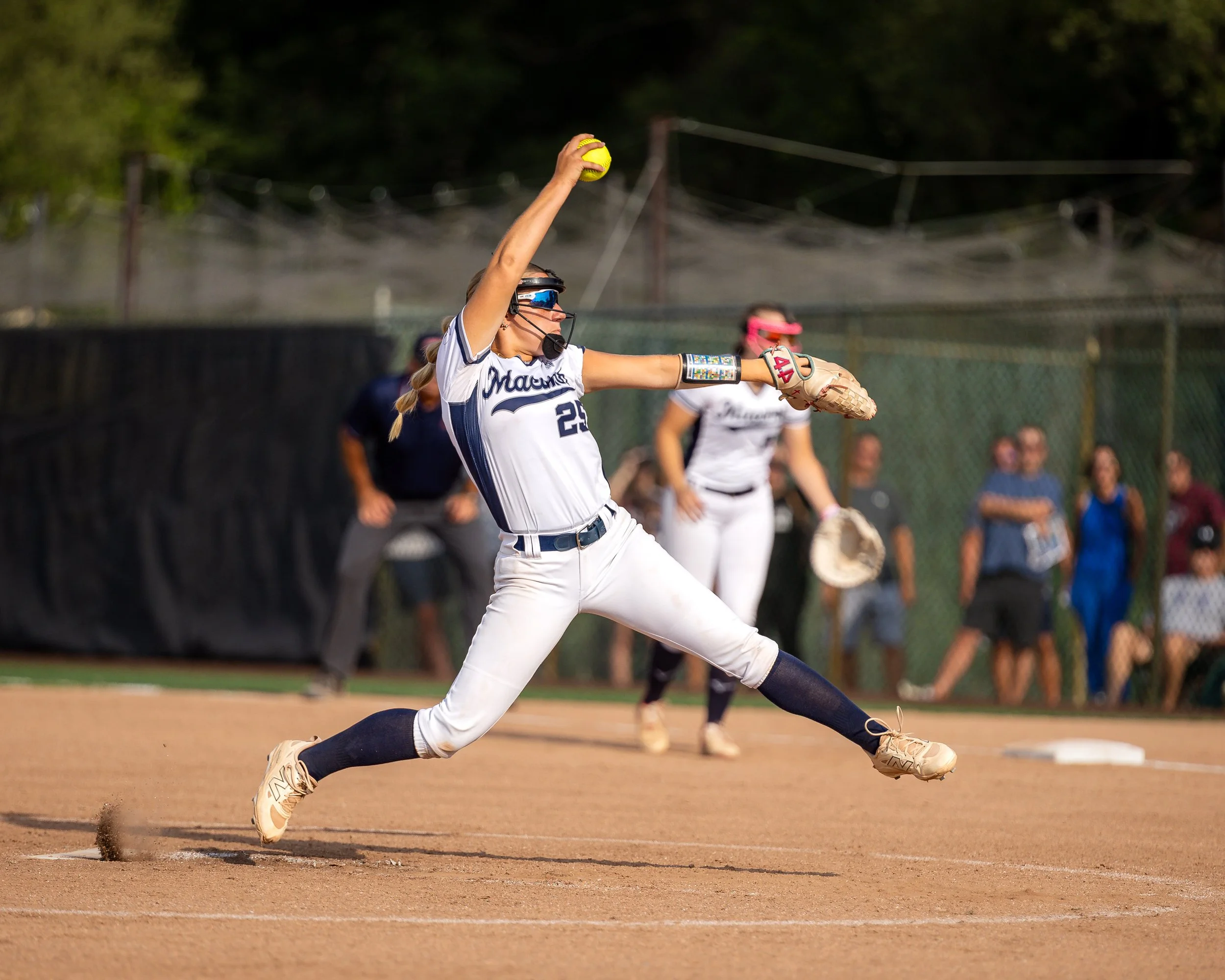 Female softball player in a white and navy uniform, pitching a ball on the field, with spectators in the background.