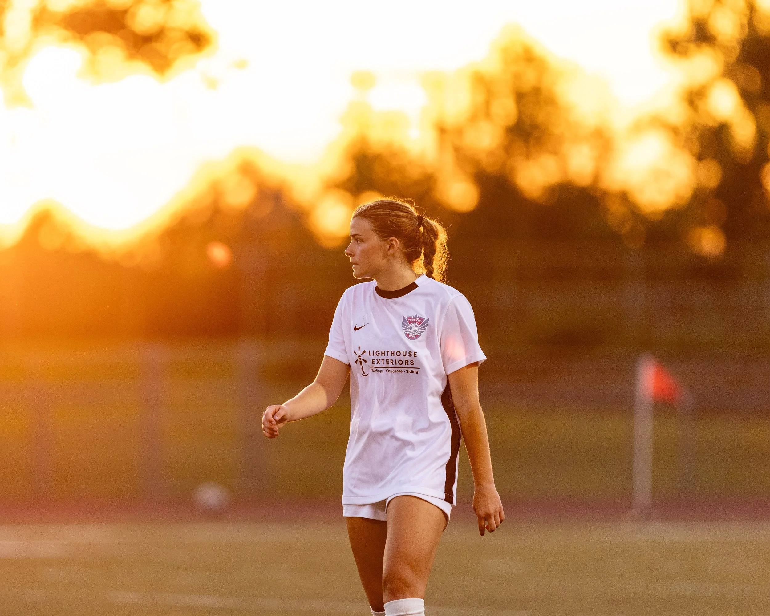 A female soccer player in a white jersey walking on the field during sunset.