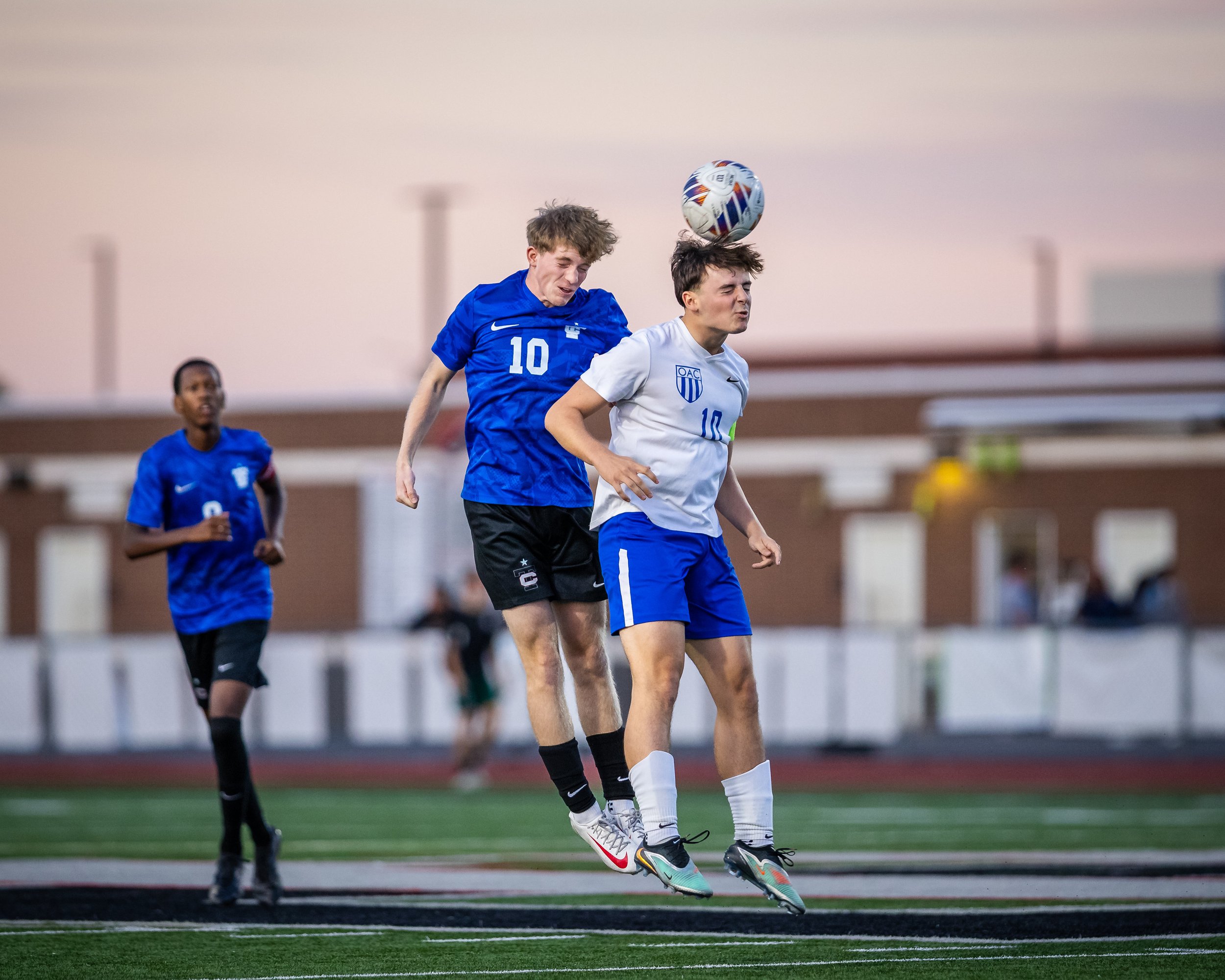 Two soccer players, one in a white jersey and the other in a blue jersey, jump to head the ball during a match on a field, with a third player running in the background.