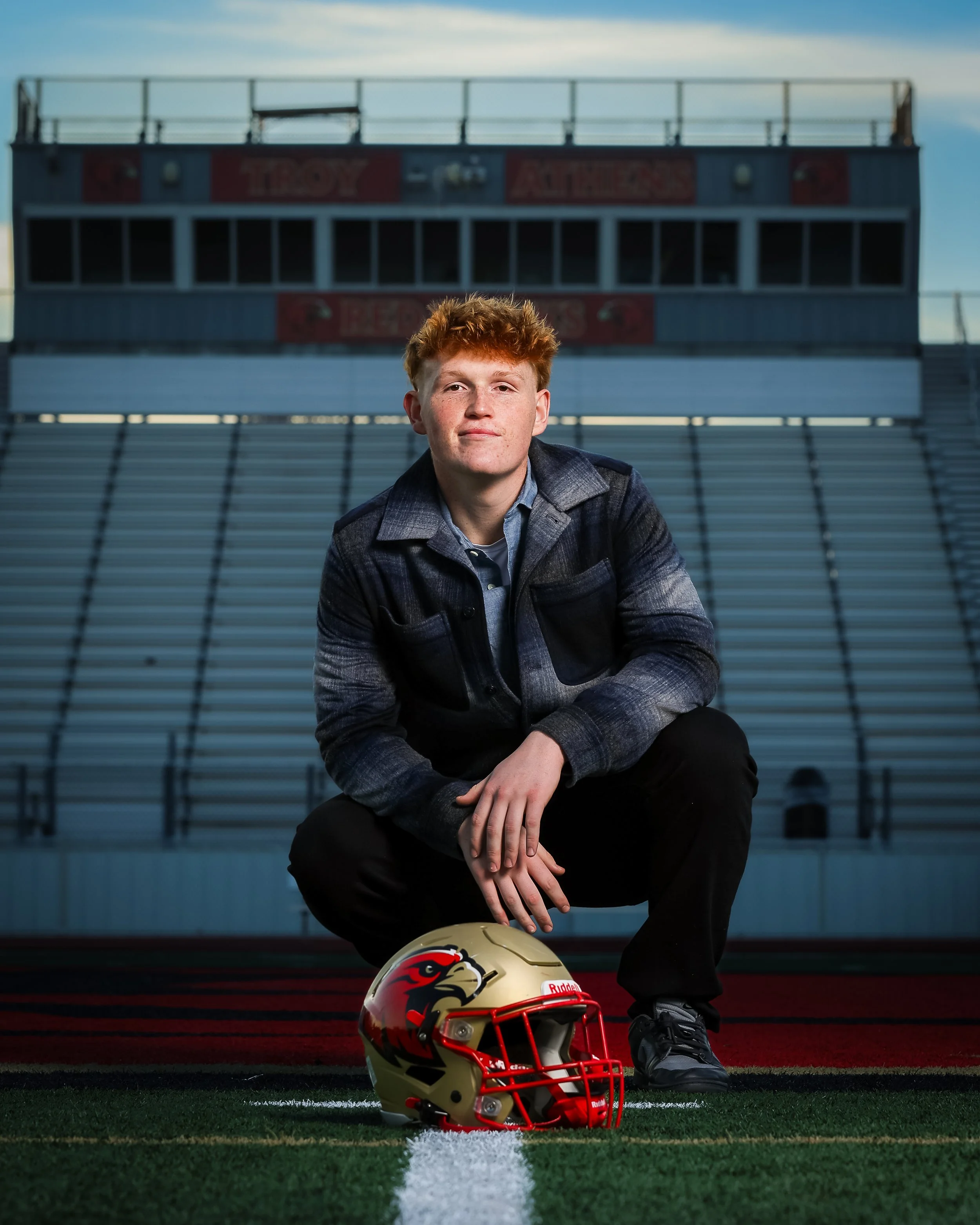 A young man with red hair crouching behind an American football helmet with a falcon logo on the field of a stadium, with empty bleachers and a press box in the background.