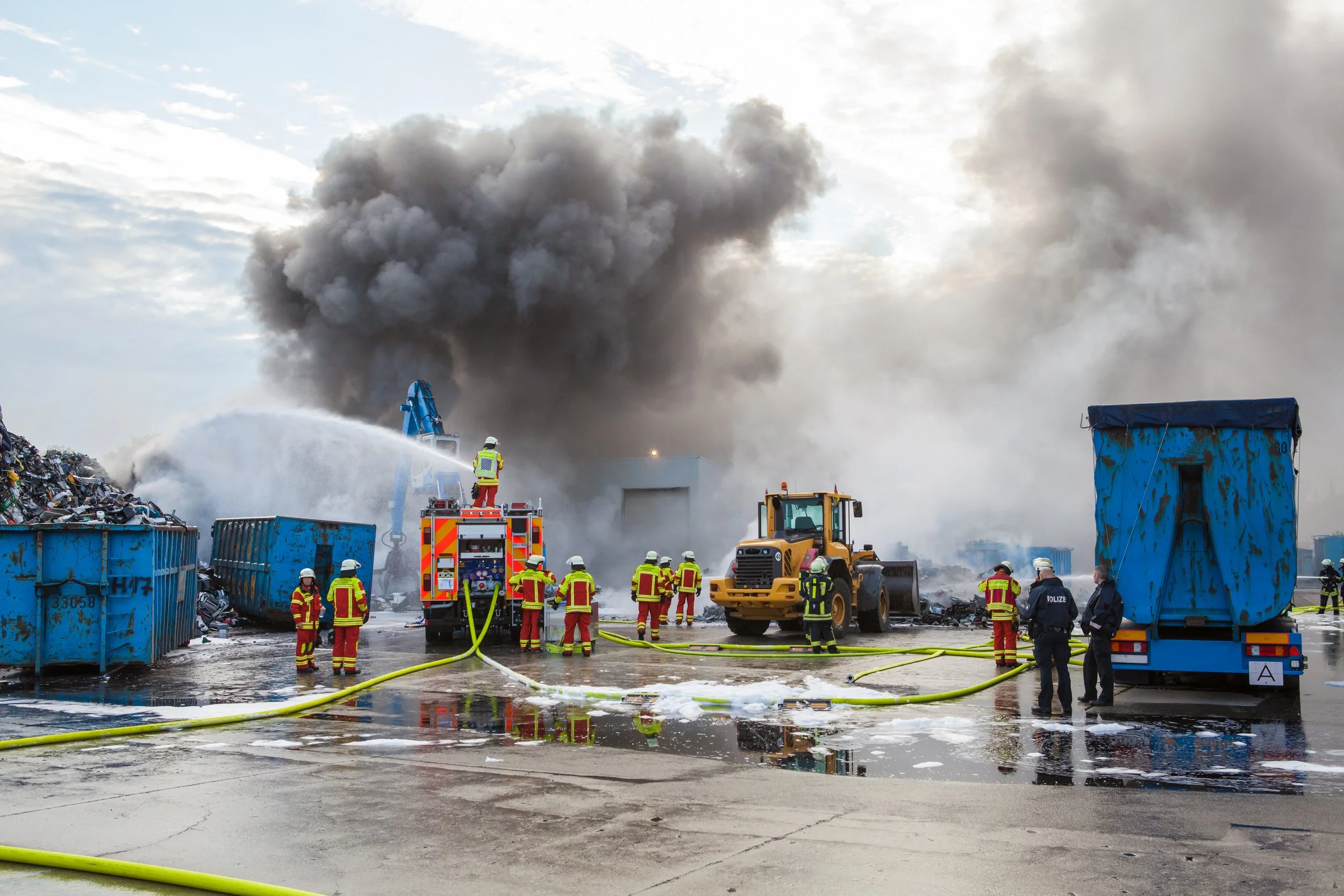 Firefighters spraying a smoking site beside blue containers and a loader