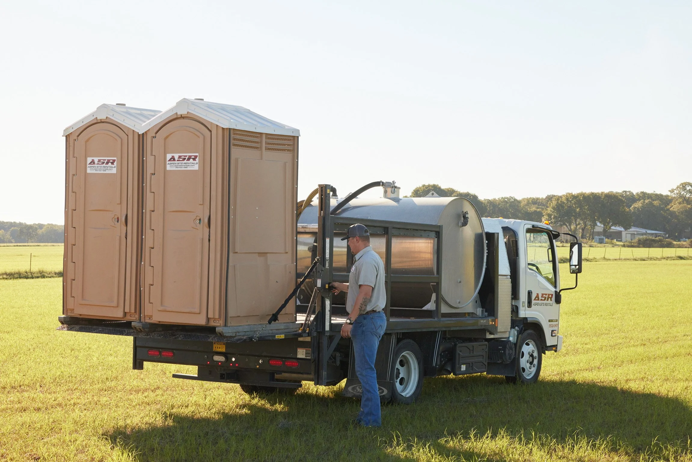 Two portable toilets loaded on a truck bed beside a stainless tank, with a man standing by