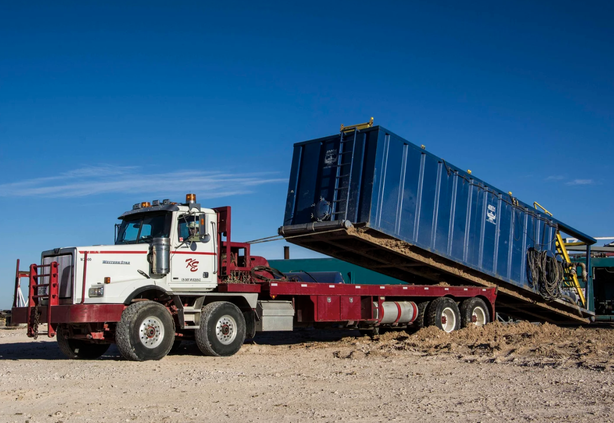 Truck tipping a blue roll-off container onto a dirt lot