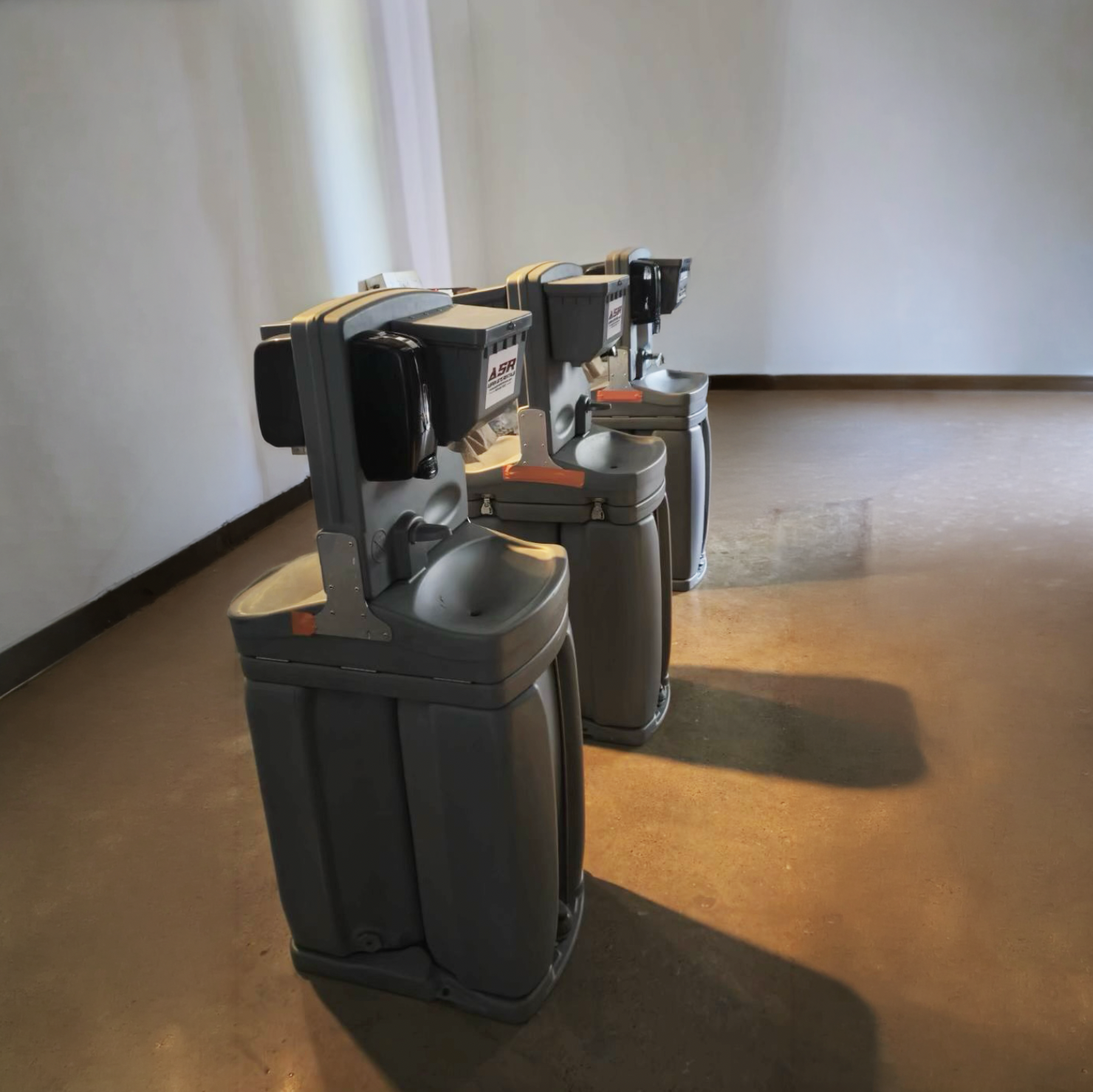 Three gray portable hand-wash stations lined up on a concrete floor
