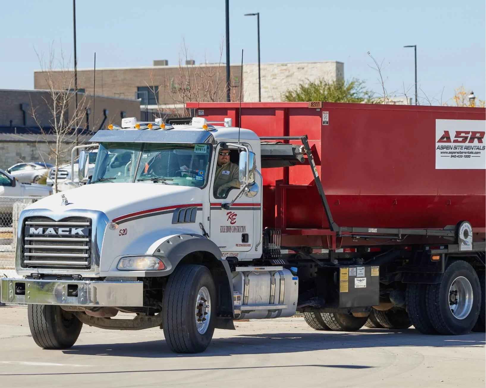 White Mack truck carrying a red roll-off dumpster in a parking lot