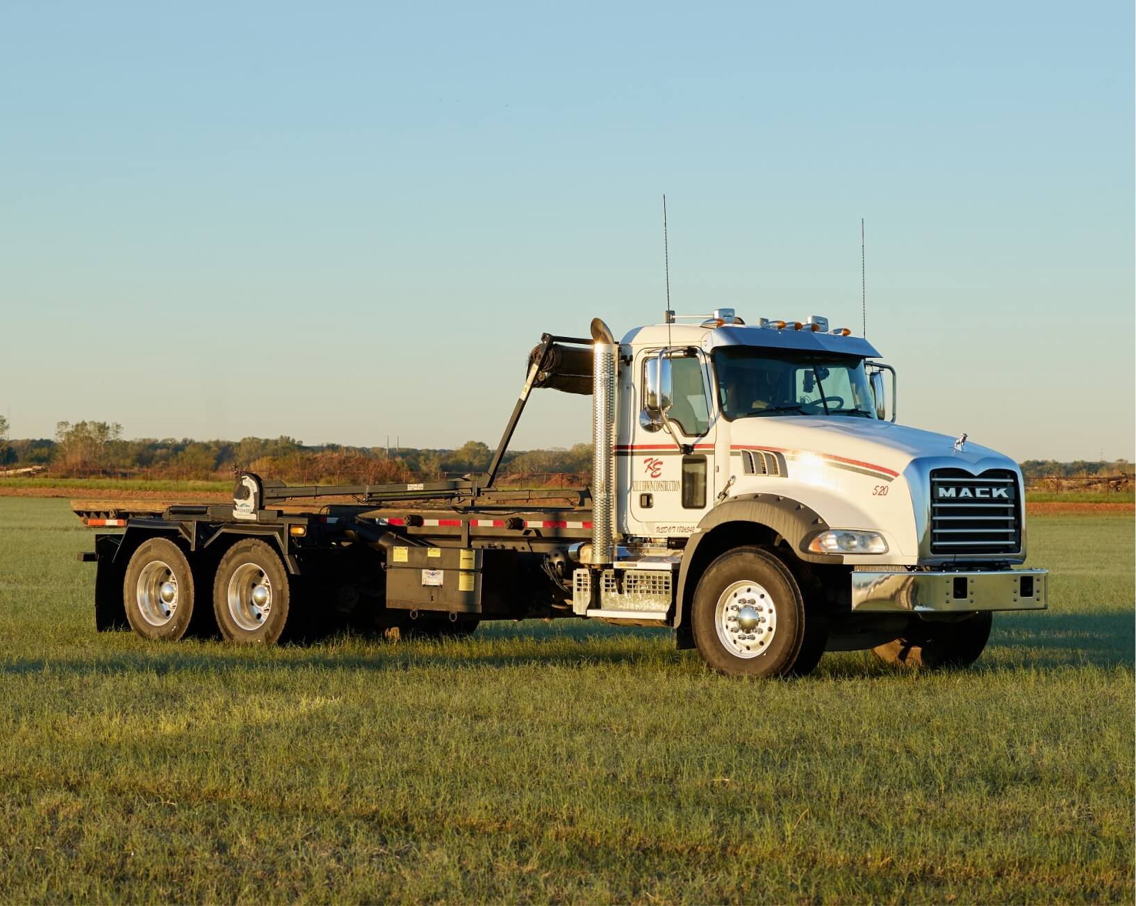 White Mack roll-off truck parked in a grass field at sunset