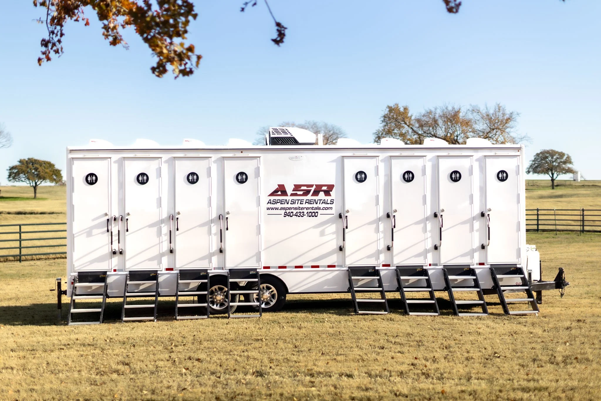 White luxury restroom trailer with eight numbered doors parked on grass by a pasture fence