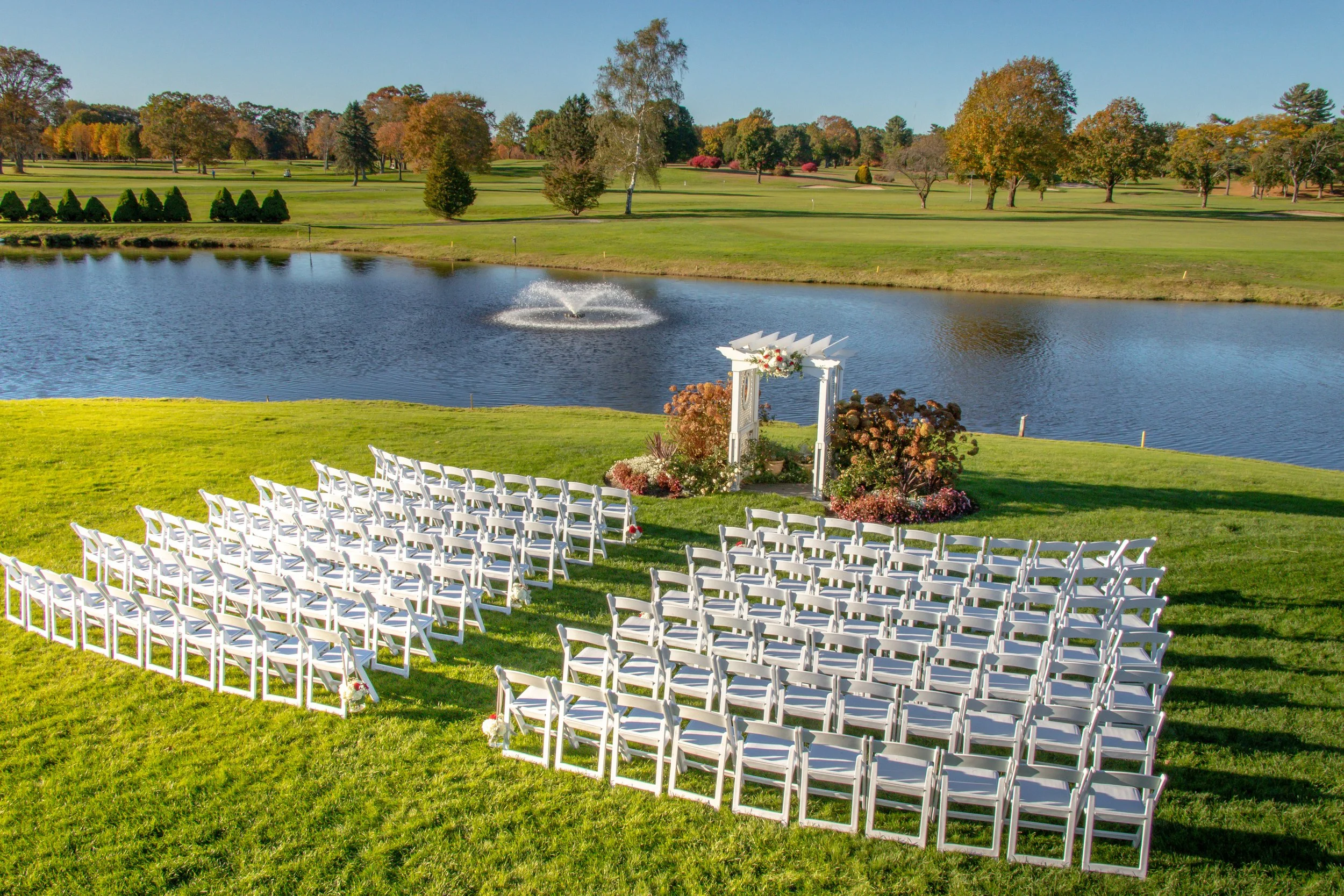 Outdoor wedding ceremony with white chairs facing an arbor by a pond