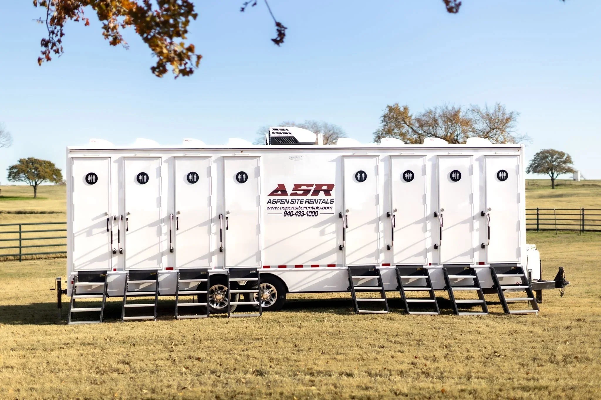 White luxury restroom trailer with eight numbered doors parked on grass by a pasture fence