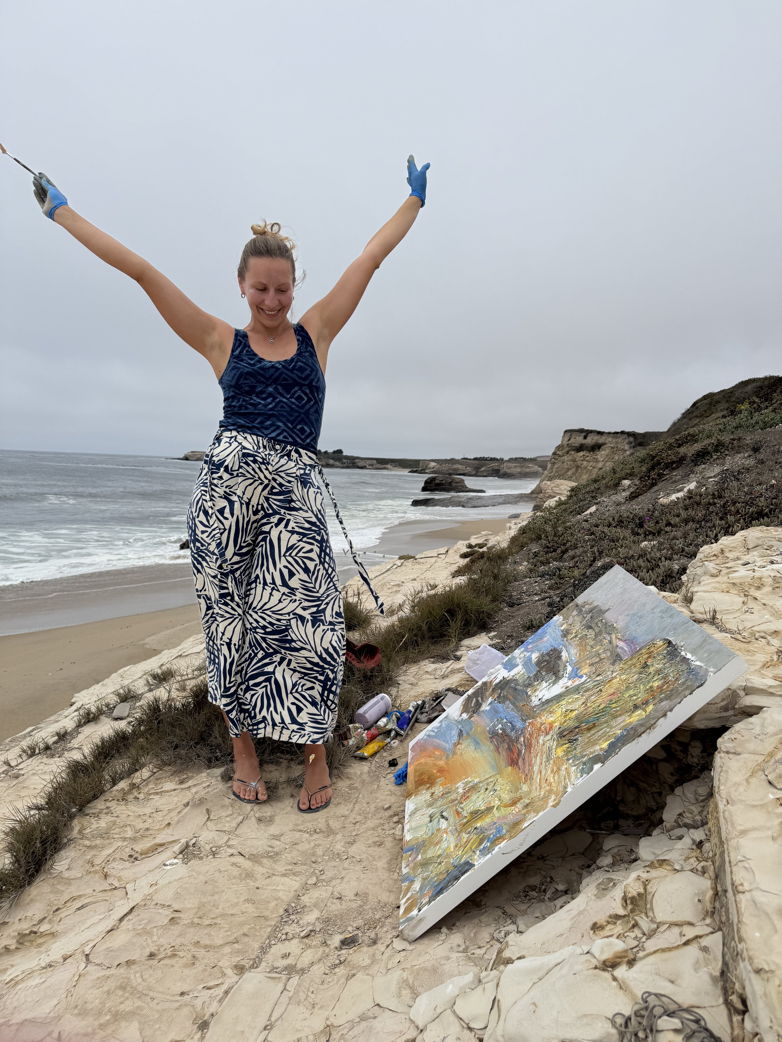 A woman wearing a blue patterned dress and sandals, smiling with arms raised, on a rocky beach with cliffs and the ocean in the background. An abstract painting and art supplies are on the ground beside her.