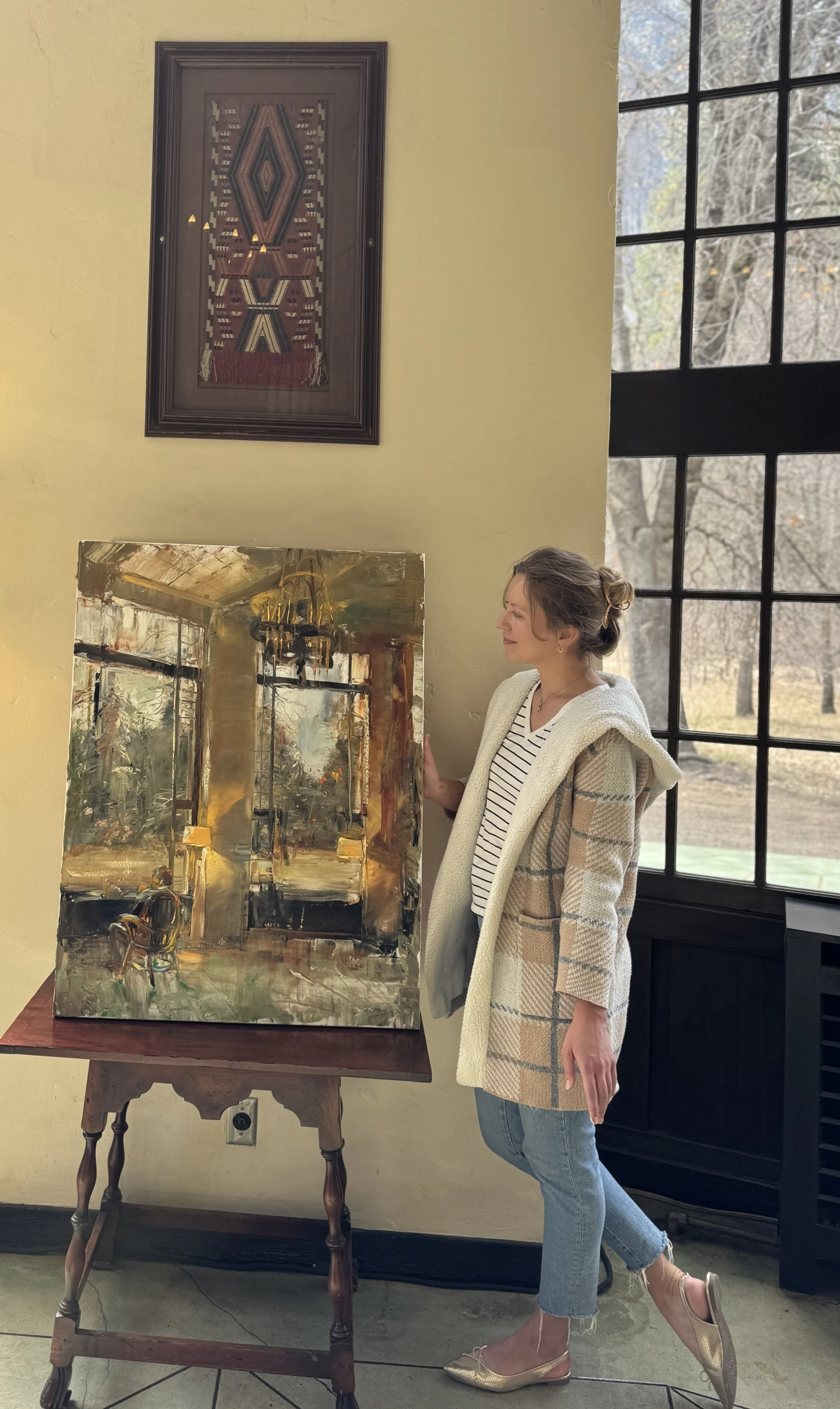 A woman with her hair in a bun, wearing a white and gray plaid coat, striped shirt, and jeans, is standing next to a framed painting on a wooden table, inside a room with large windows and a yellow wall.