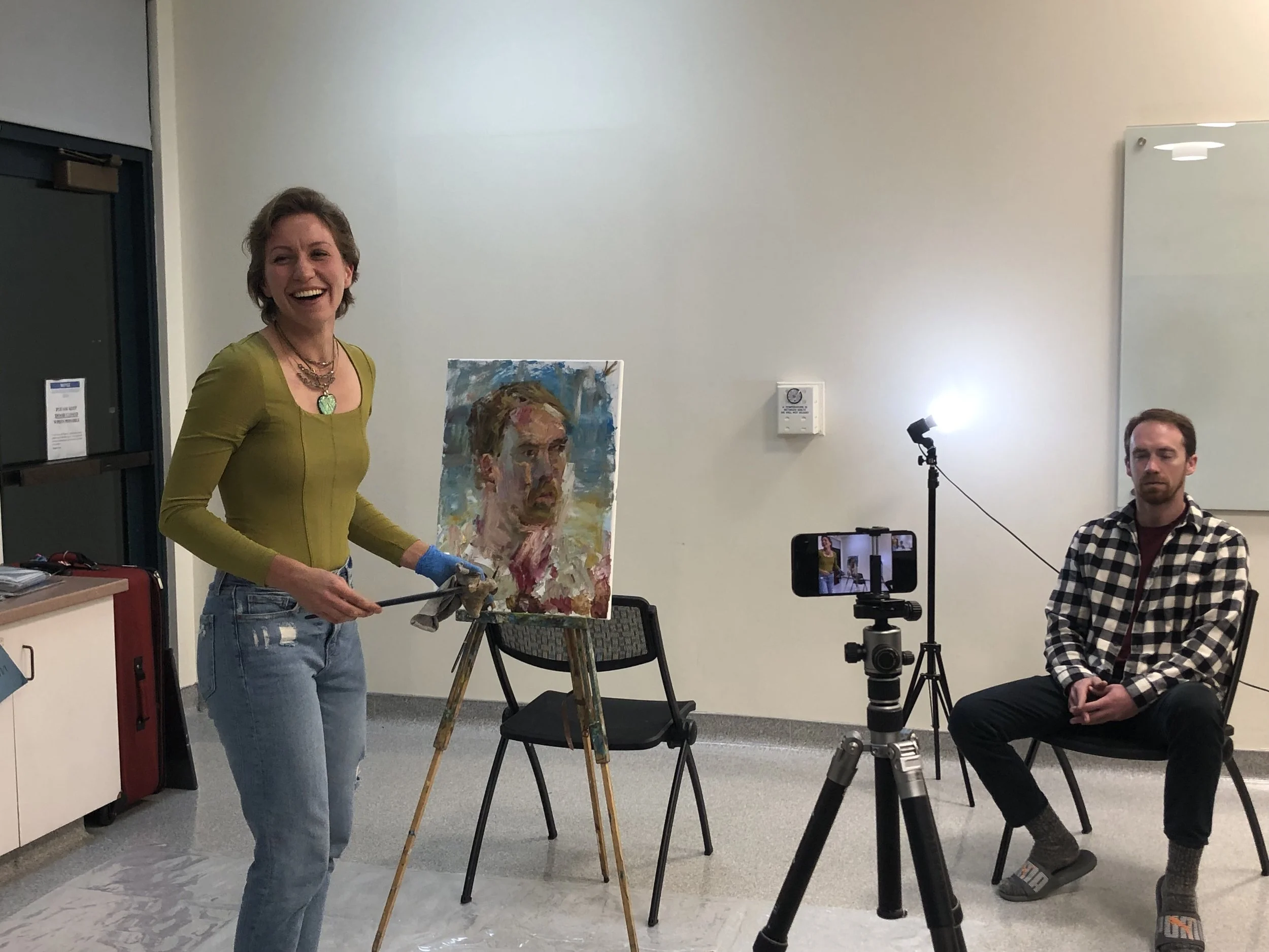 A woman with short hair in a yellow top and jeans is painting a portrait of a man. She is smiling and using tools for painting. There is a man in a checkered shirt sitting nearby, and a camera on a tripod is pointing toward the woman. The painting on the easel appears to be an abstract or impressionistic portrait of a man.