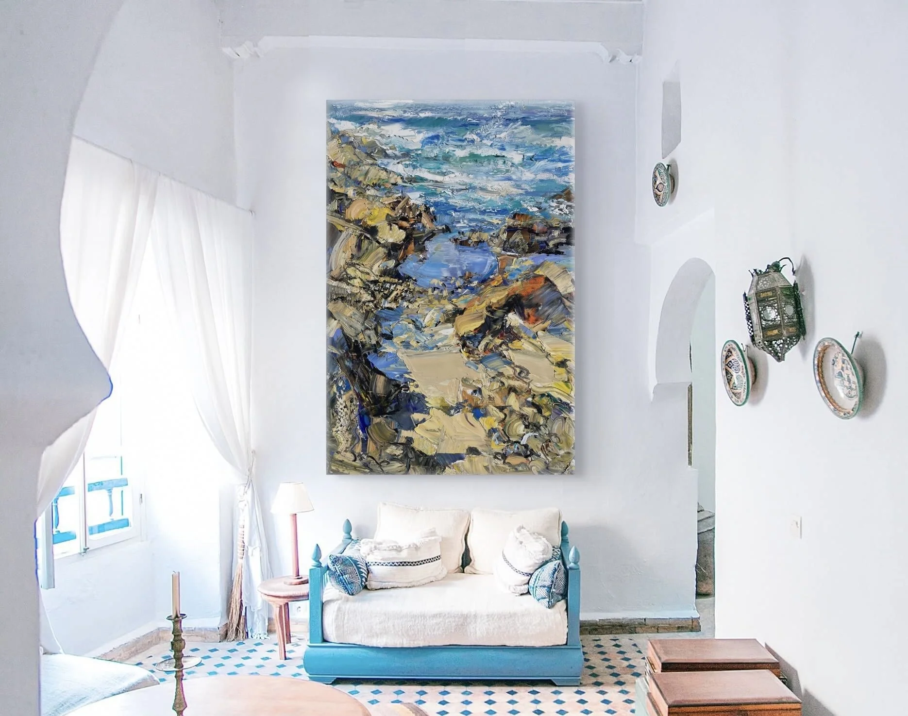 A bright, coastal-themed living room with white walls and accents of blue. A large, colorful painting of a seaside landscape hangs above a white wooden sofa with cushions. There are decorative plates on the walls, and a vintage lantern on a shelf. A patterned tile floor is partially visible.