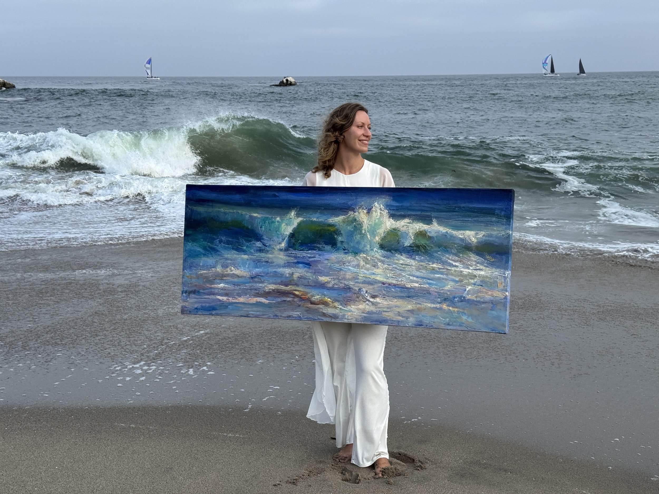 A woman standing barefoot on the beach holding a large painting of ocean waves, with the sea and sailboats in the background.