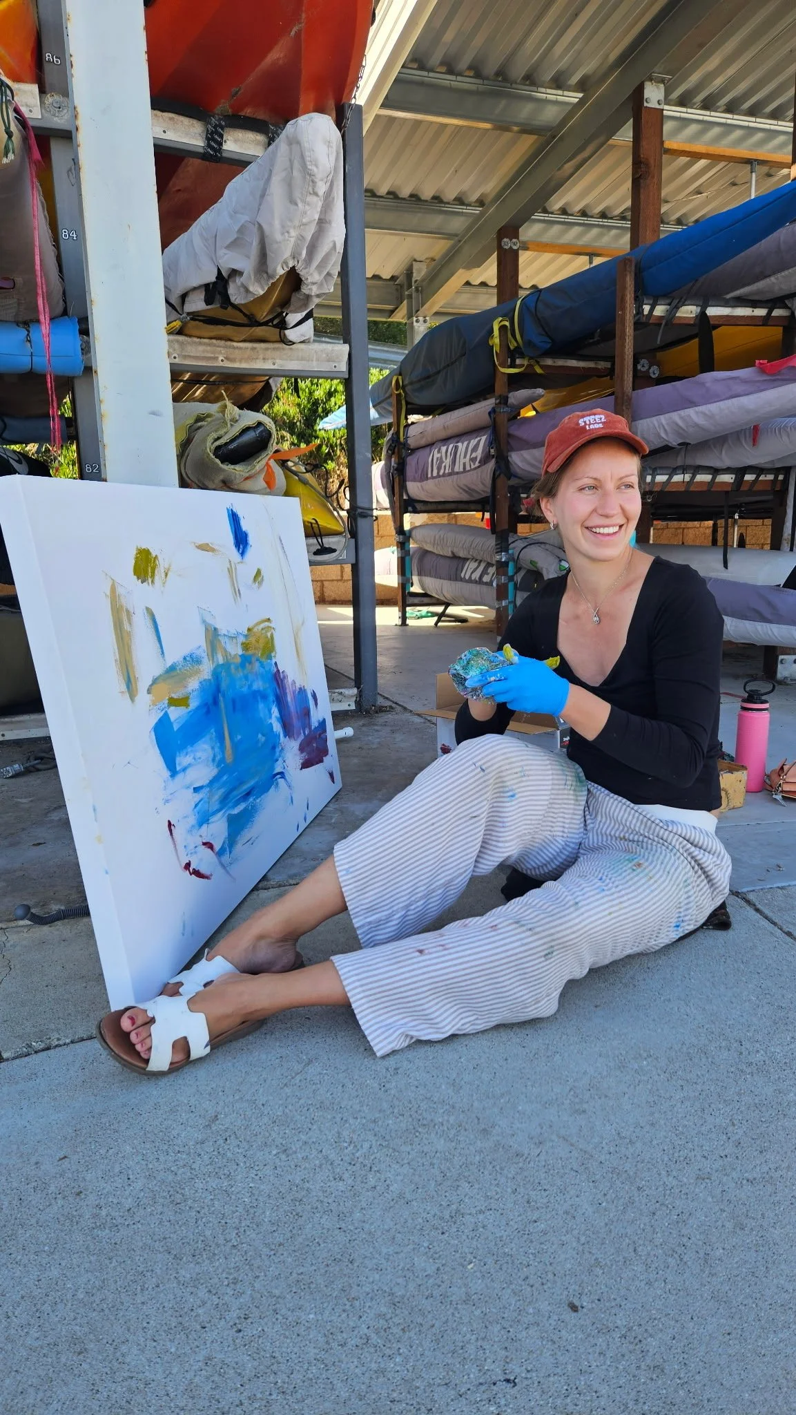A woman sitting on the ground, smiling, holding a palette in her hands. She is wearing a black top, striped pants, white sandals, and a red cap. There is a large, colorful abstract painting on the ground in front of her. Behind her are shelves with life jackets and other gear under a metal roof.