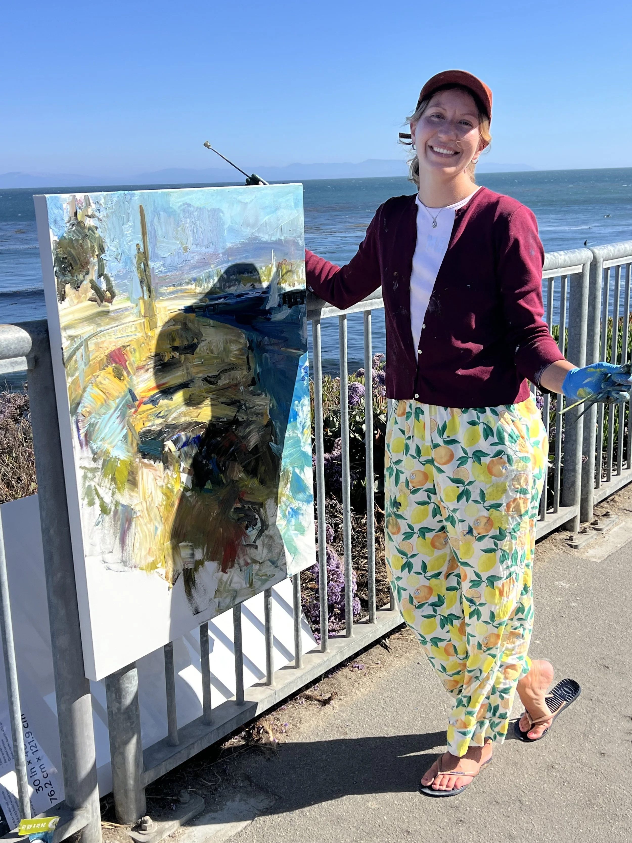 A woman with light skin, wearing a red cap, white shirt, maroon cardigan, and lemon-patterned yellow and white pants, is smiling while painting an outdoor scene near the ocean. She is standing on a sidewalk, holding a paintbrush, with a colorful abstract painting on an easel beside her. The ocean and distant mountains can be seen in the background under a clear blue sky.