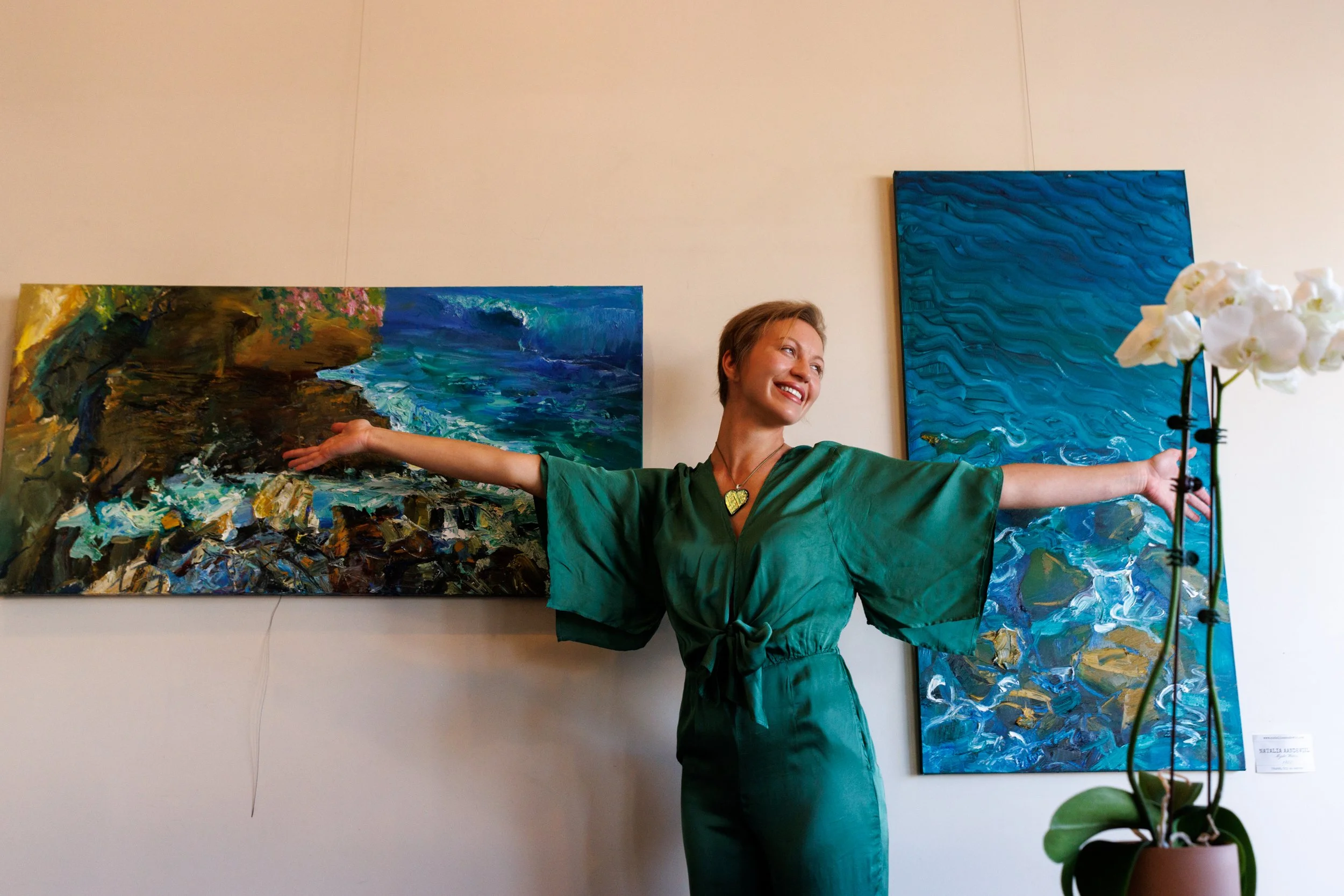 A woman with short hair and fair skin stands in front of two vibrant abstract paintings, smiling with her arms outstretched. She wears a green wrap dress and a necklace with a heart-shaped pendant. A white orchid plant is visible in the foreground.