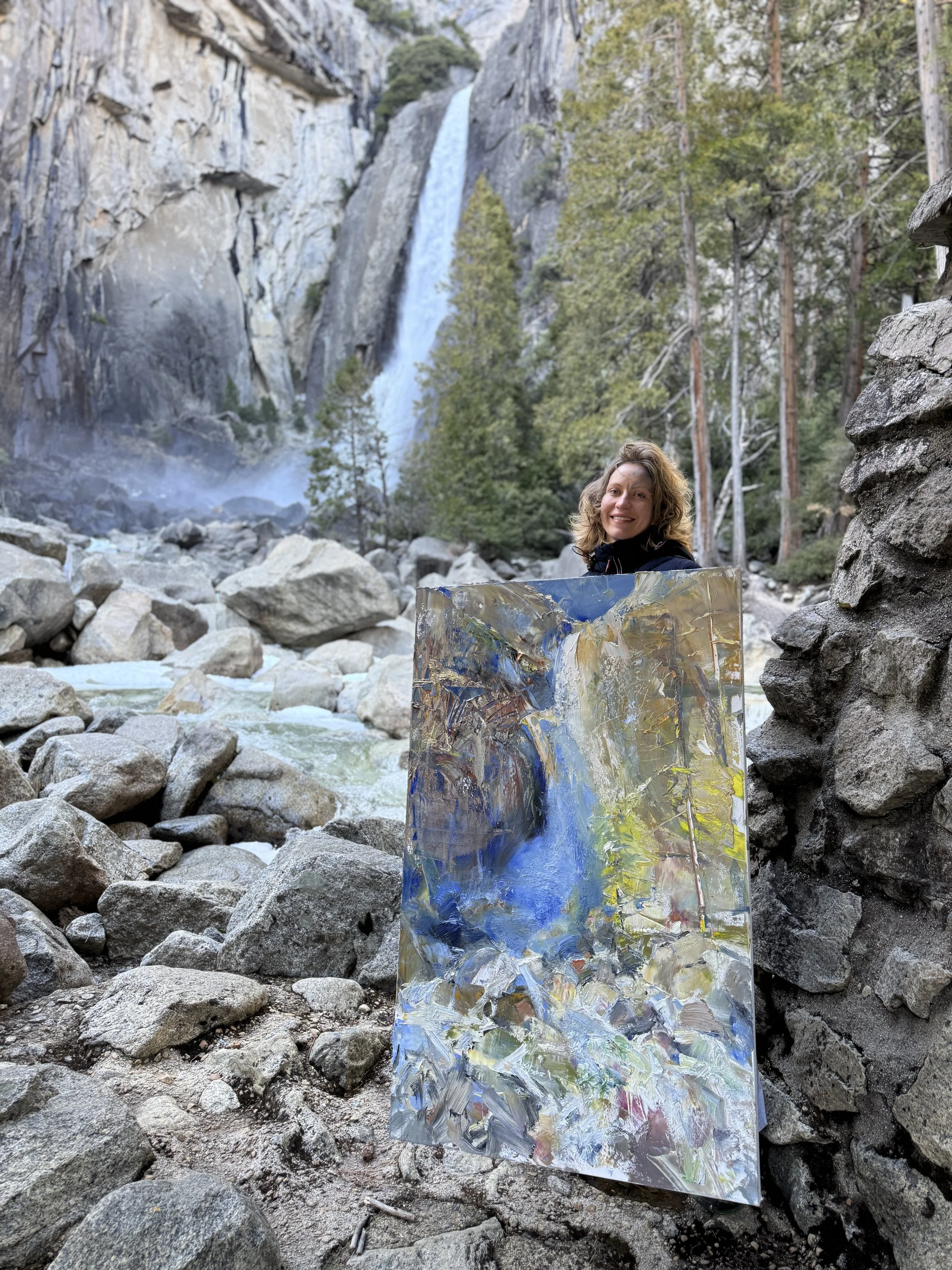 A woman holding a colorful abstract painting outdoors in front of rocks, trees, and a waterfall in a mountainous area.