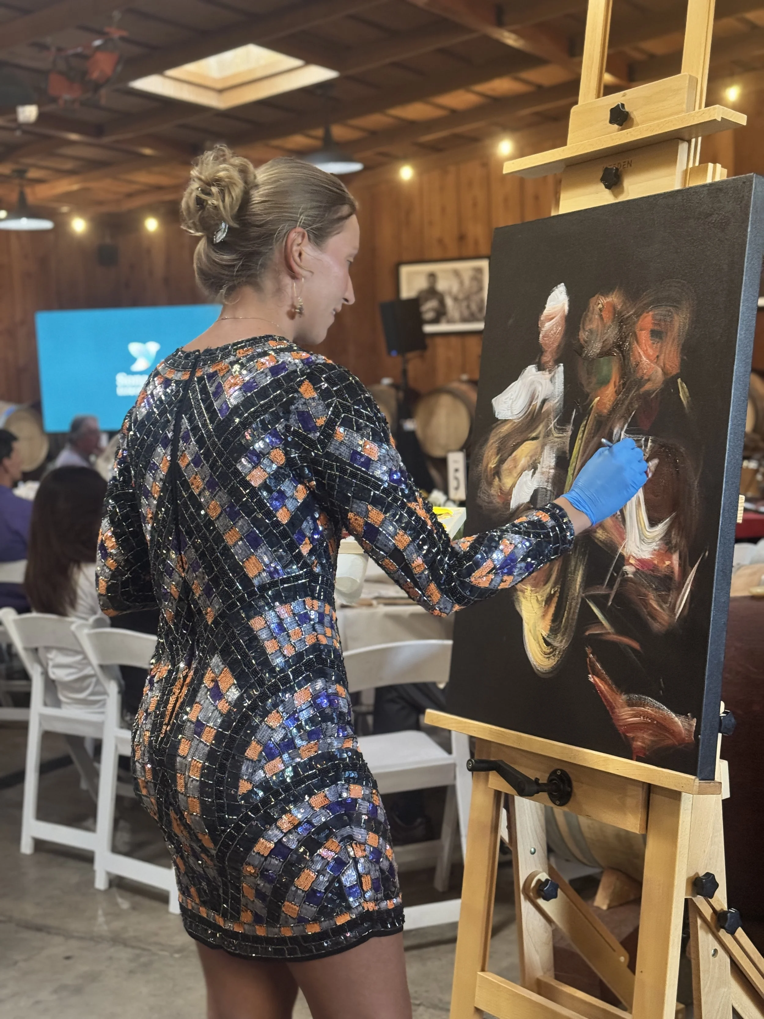 A woman with her hair styled in an updo is painting an abstract piece on a canvas at an art event held in a wooden venue. She is wearing a patterned dress and a blue glove on her right hand.