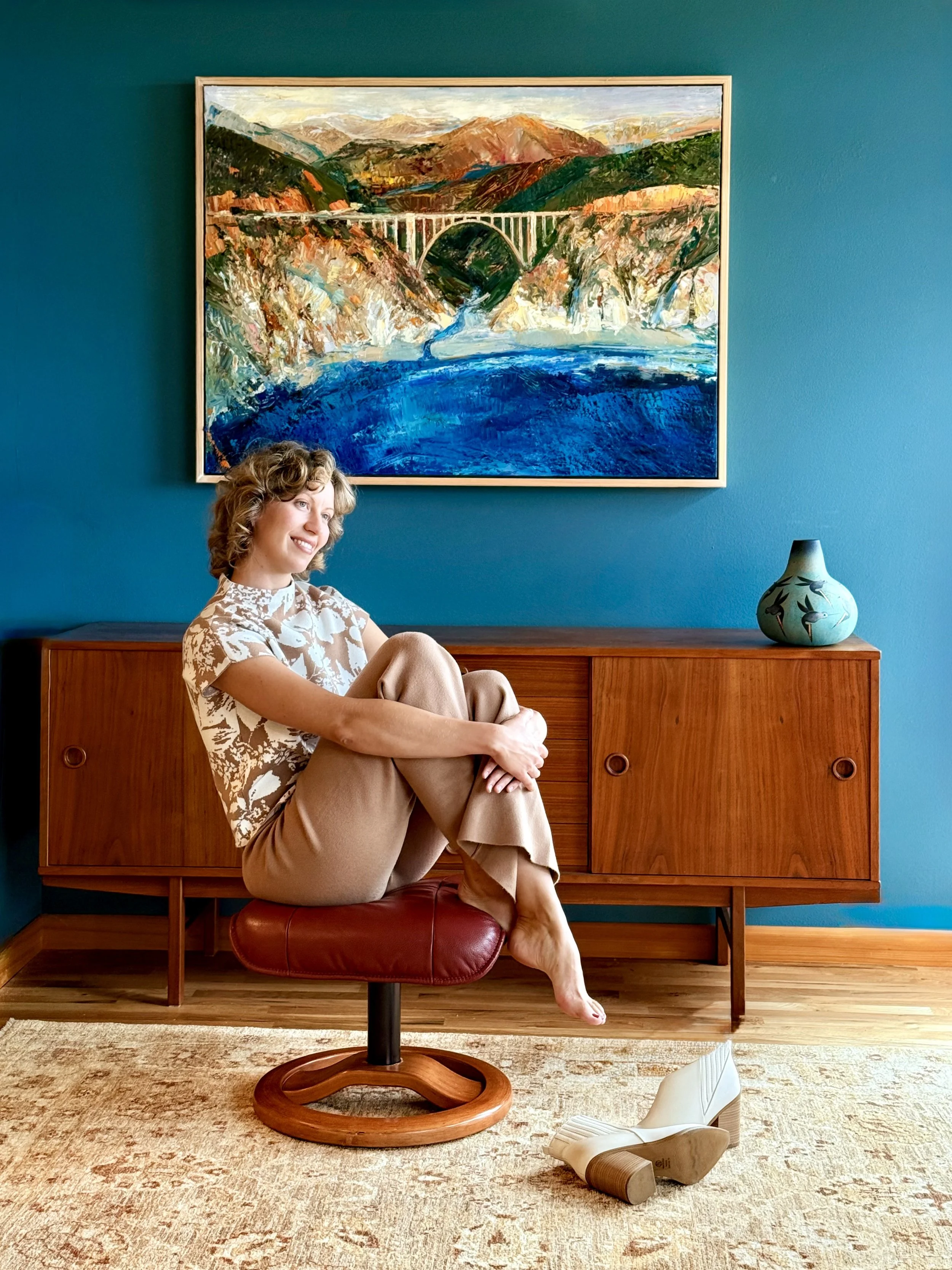 Woman sitting on a red swivel chair in front of a teal wall, with a large colorful landscape painting, a wooden sideboard, and a decorative vase on top. A white shoe is on the floor nearby.