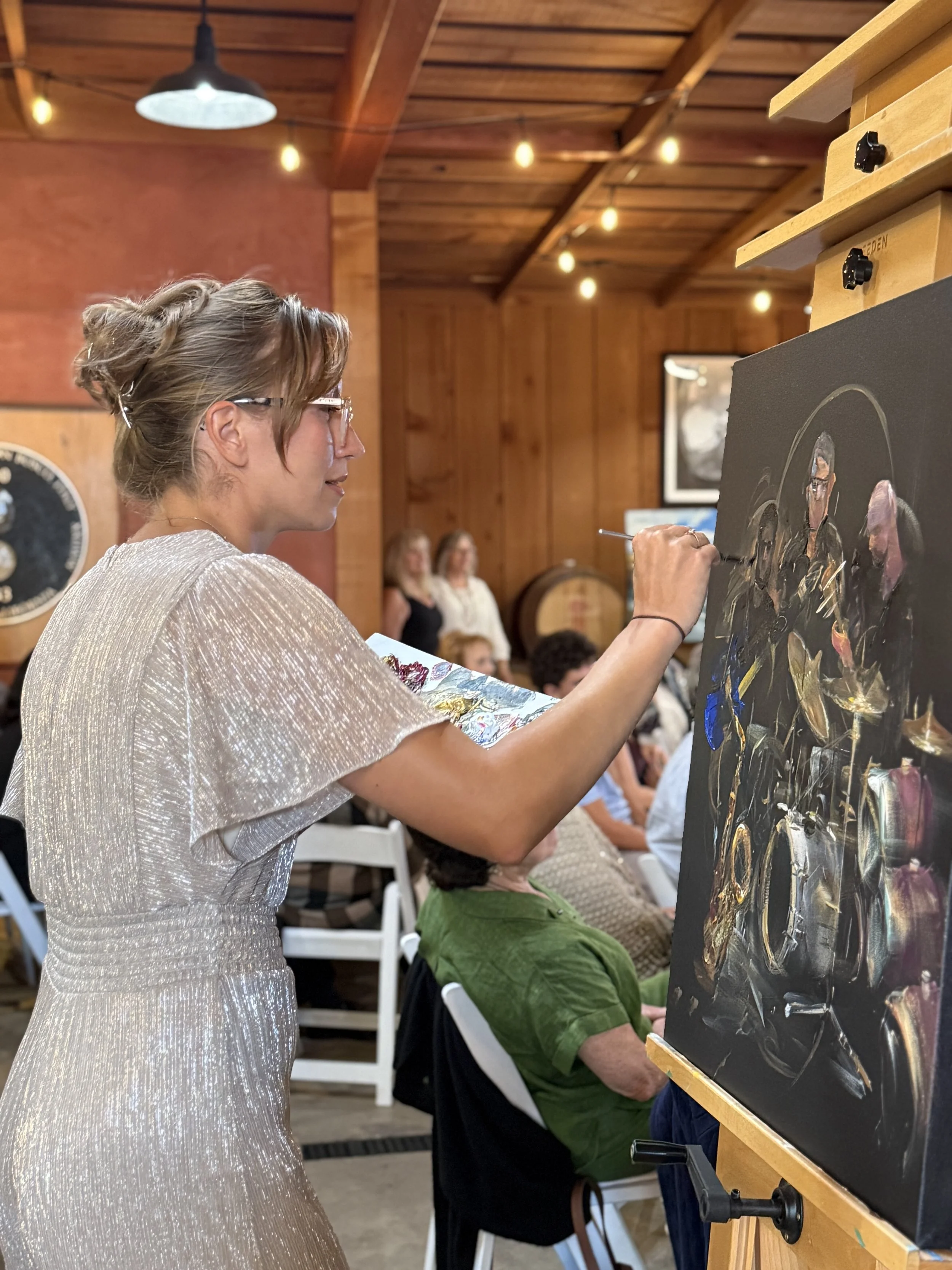 Woman with short hair and glasses painting on large canvas at art event, with audience seated in background