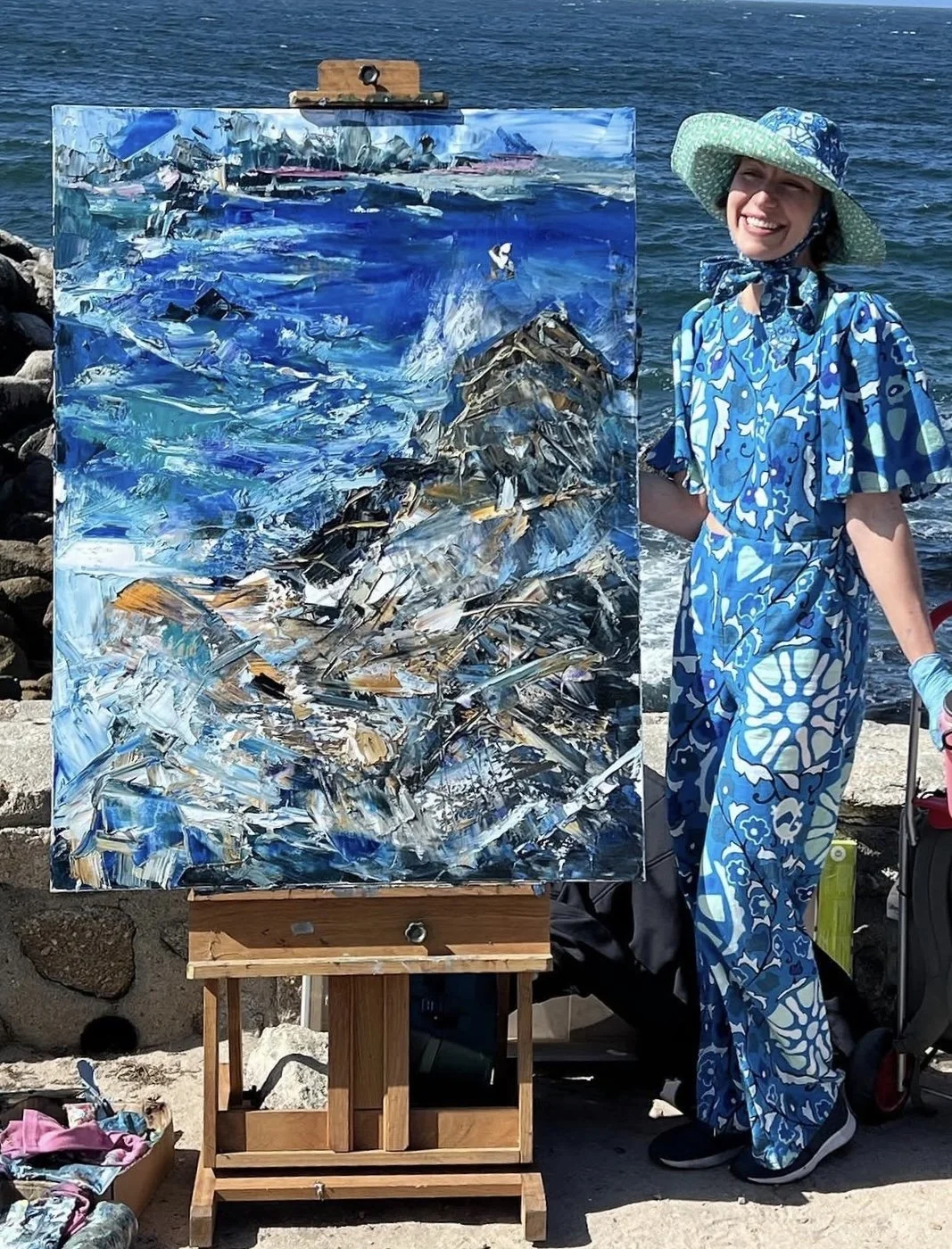 A woman in a blue floral dress and wide-brimmed hat is standing on a rocky beach, smiling, next to an abstract painting of a seascape on an easel.
