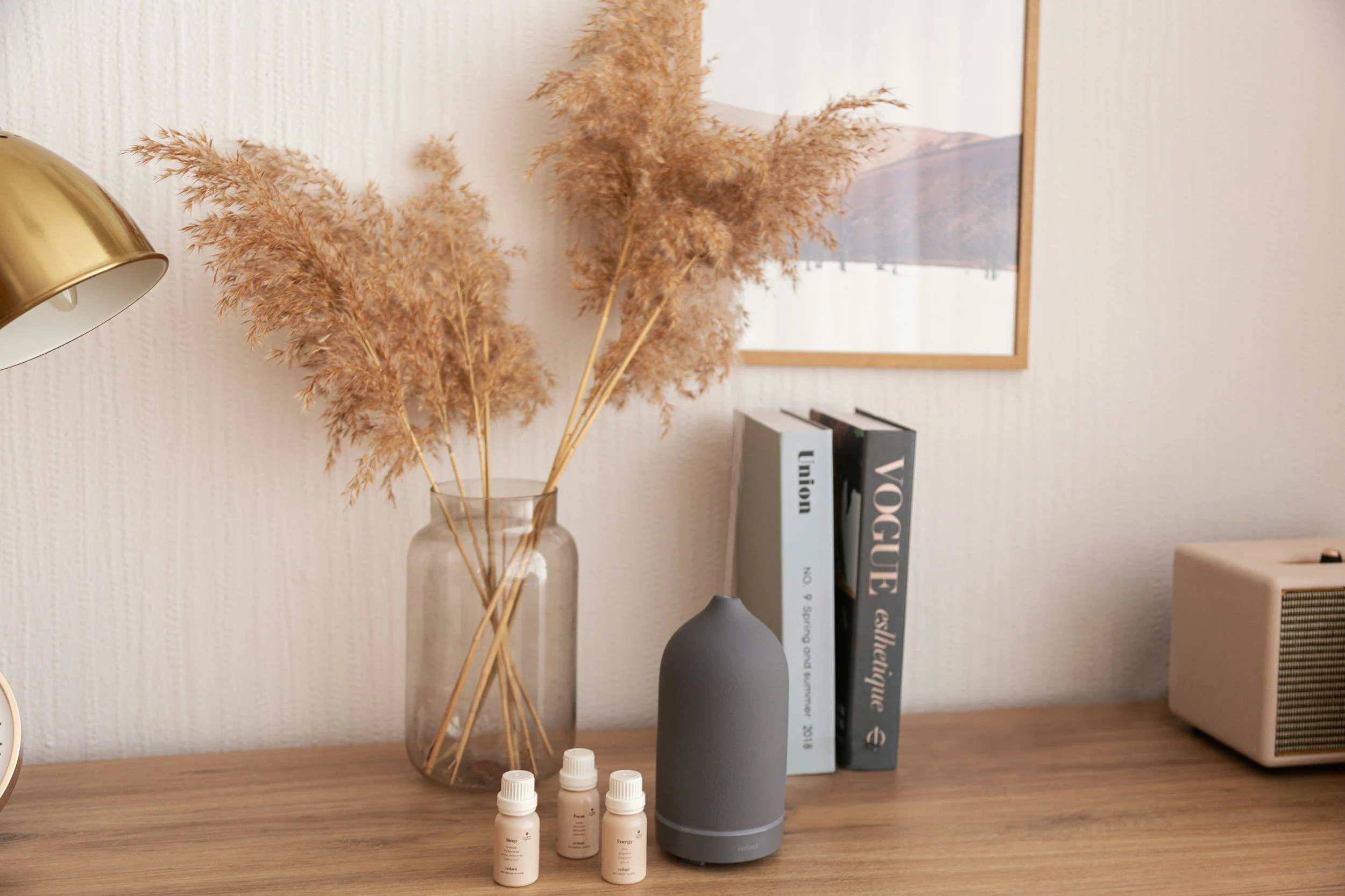 A wooden table with a gray essential oil diffuser, four small bottles of essential oils, a vase with dried pampas grass, two books, a framed wall art photography, a beige speaker, and a gold desk lamp.