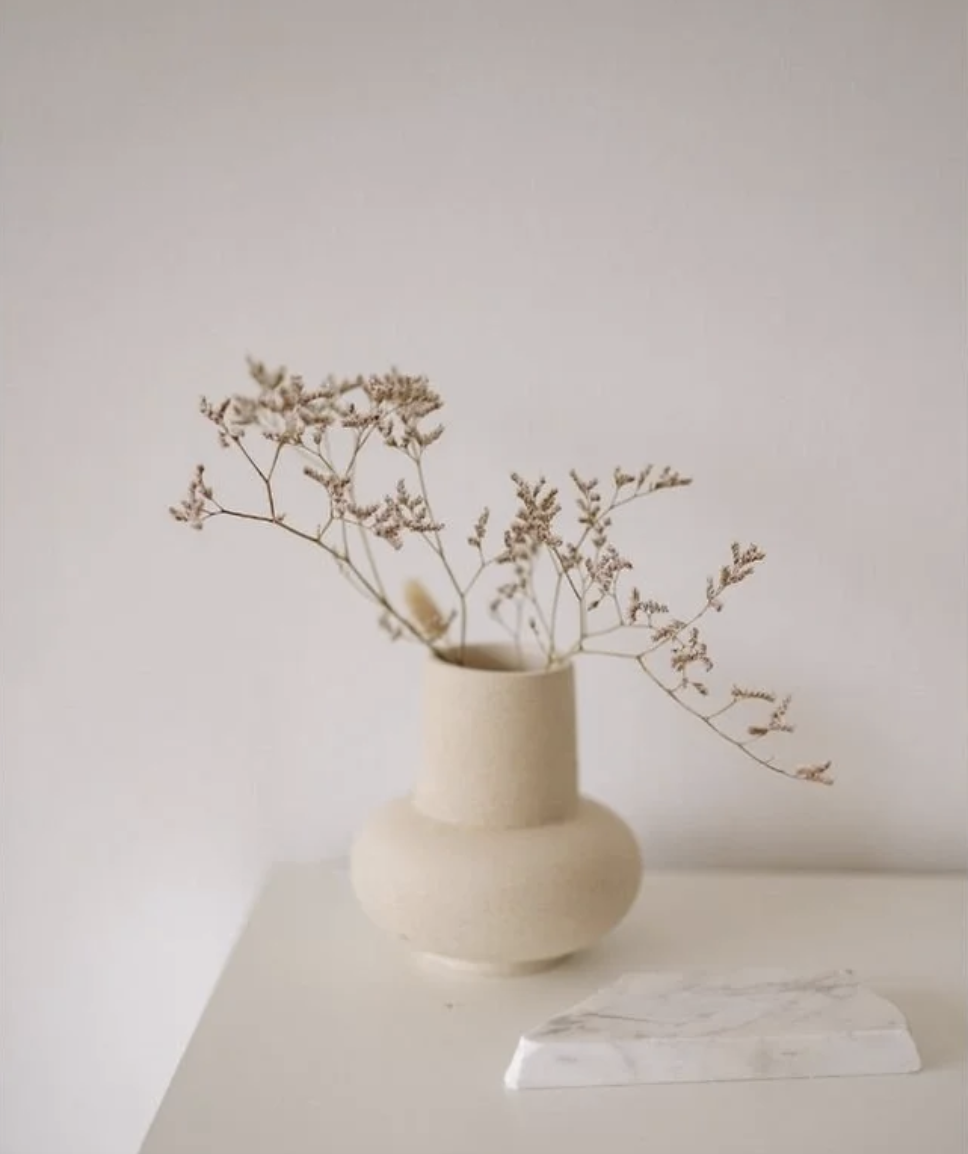 A beige vase with dried flowers on a white surface against a light background.