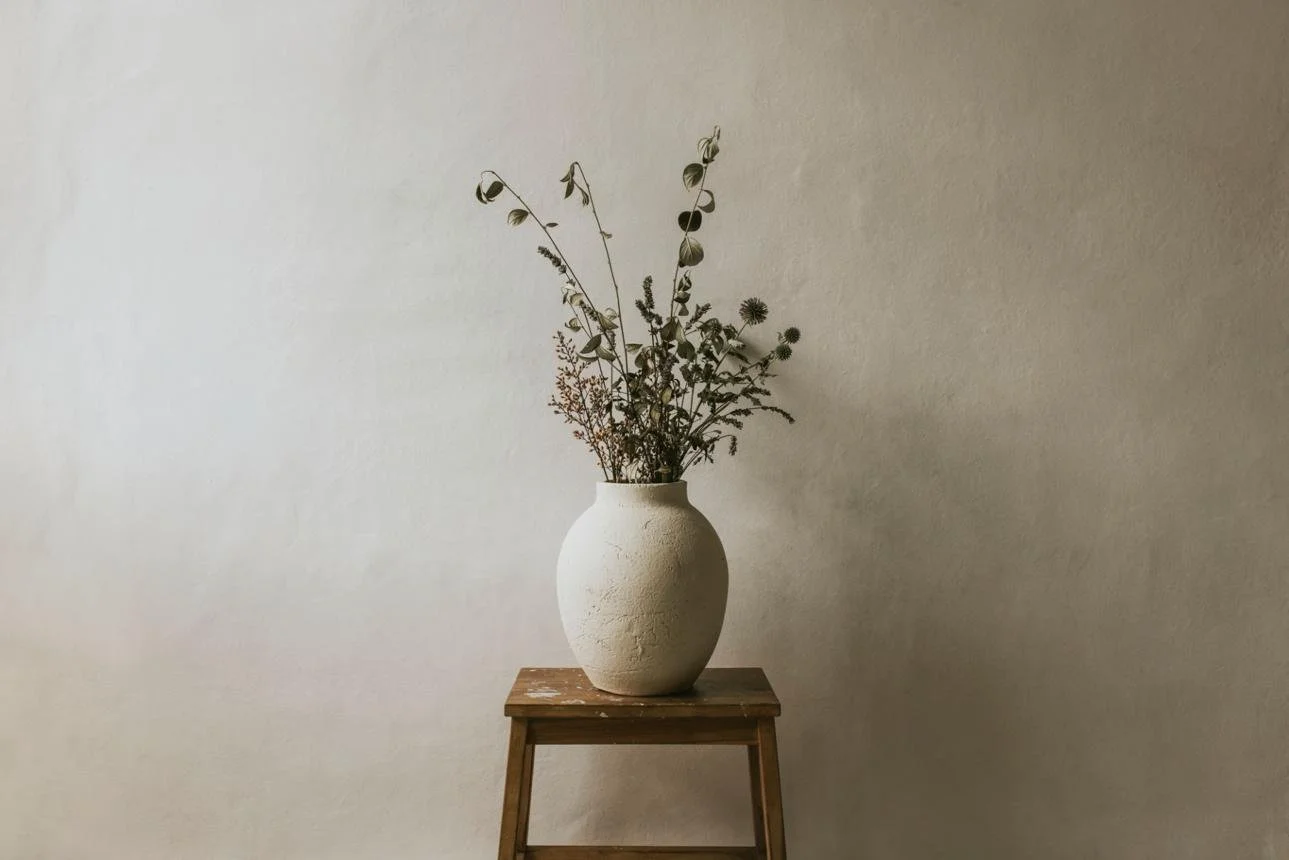 A large, round, textured white ceramic vase with dried, muted green and brown plants, placed on a small wooden stool against a plain, light-colored wall.
