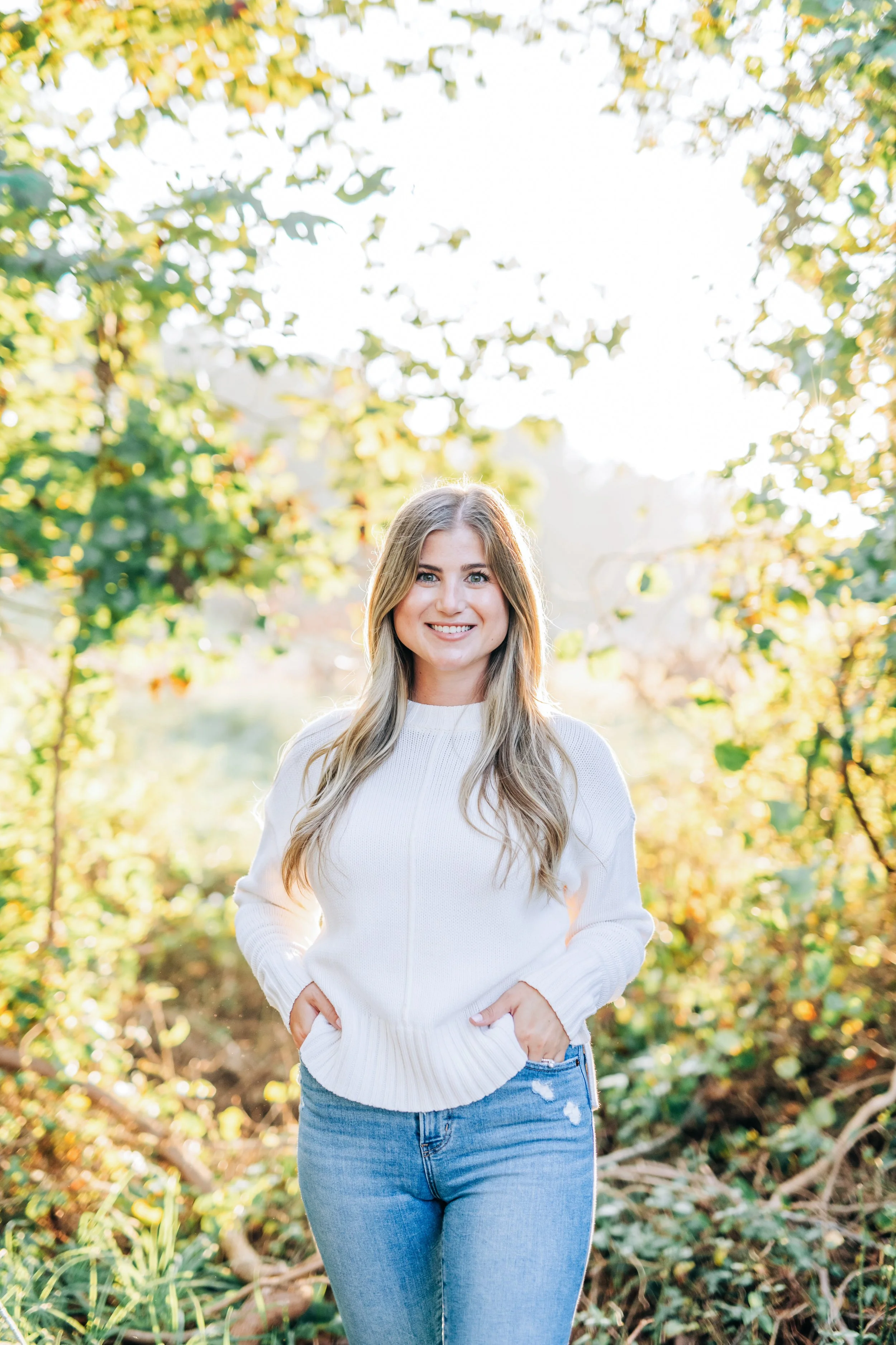 A young woman with long blonde hair wearing a white sweater and blue jeans standing outdoors in a wooded area with sunlight filtering through trees.