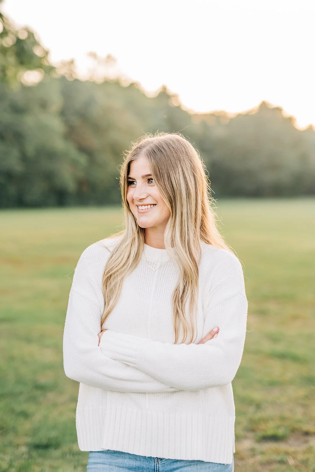A young woman with long blonde hair smiling with arms crossed outdoors in a field with green grass and trees in the background