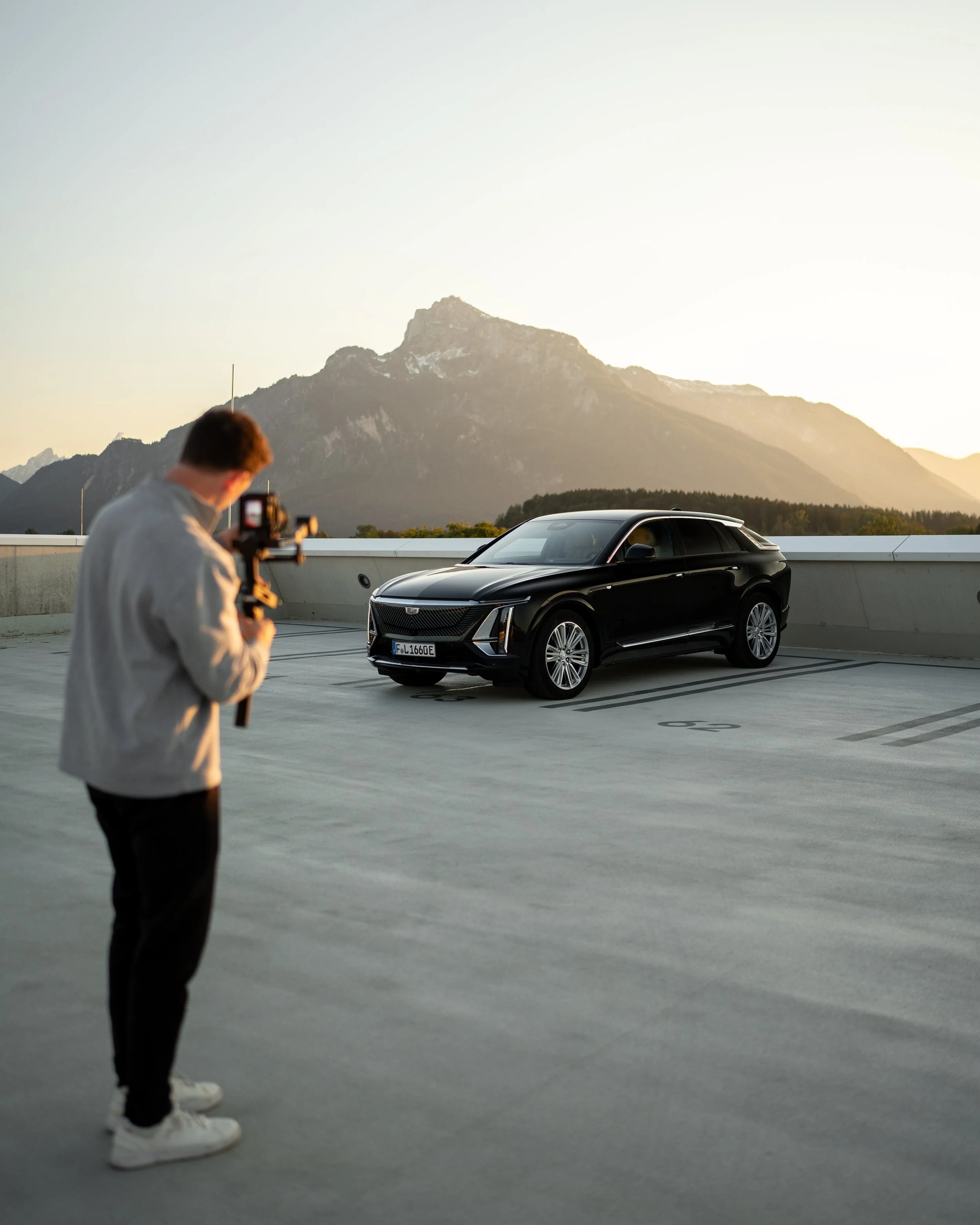 Mann, der auf einer Parkgarage steht und ein Foto mit einer Kamera macht, mit einem schwarzen Auto im Vordergrund und einer Bergkulisse im Hintergrund bei Sonnenuntergang.