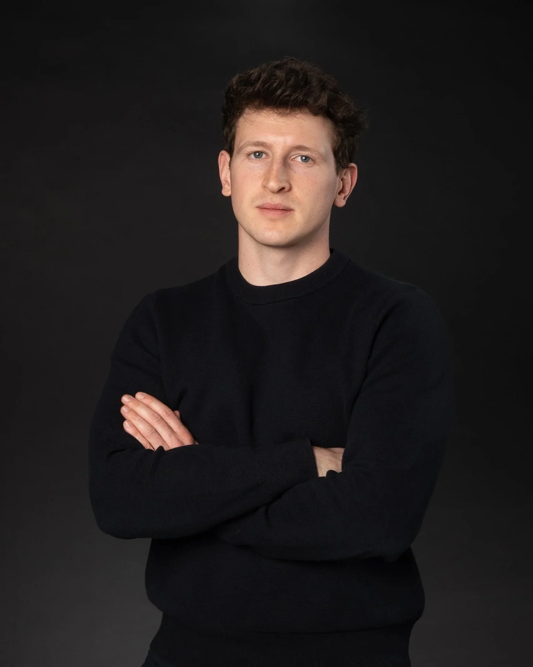 Junge Mann mit braunen, lockigen Haaren trägt einen schwarzen Pullover vor einem schwarzen Hintergrund.