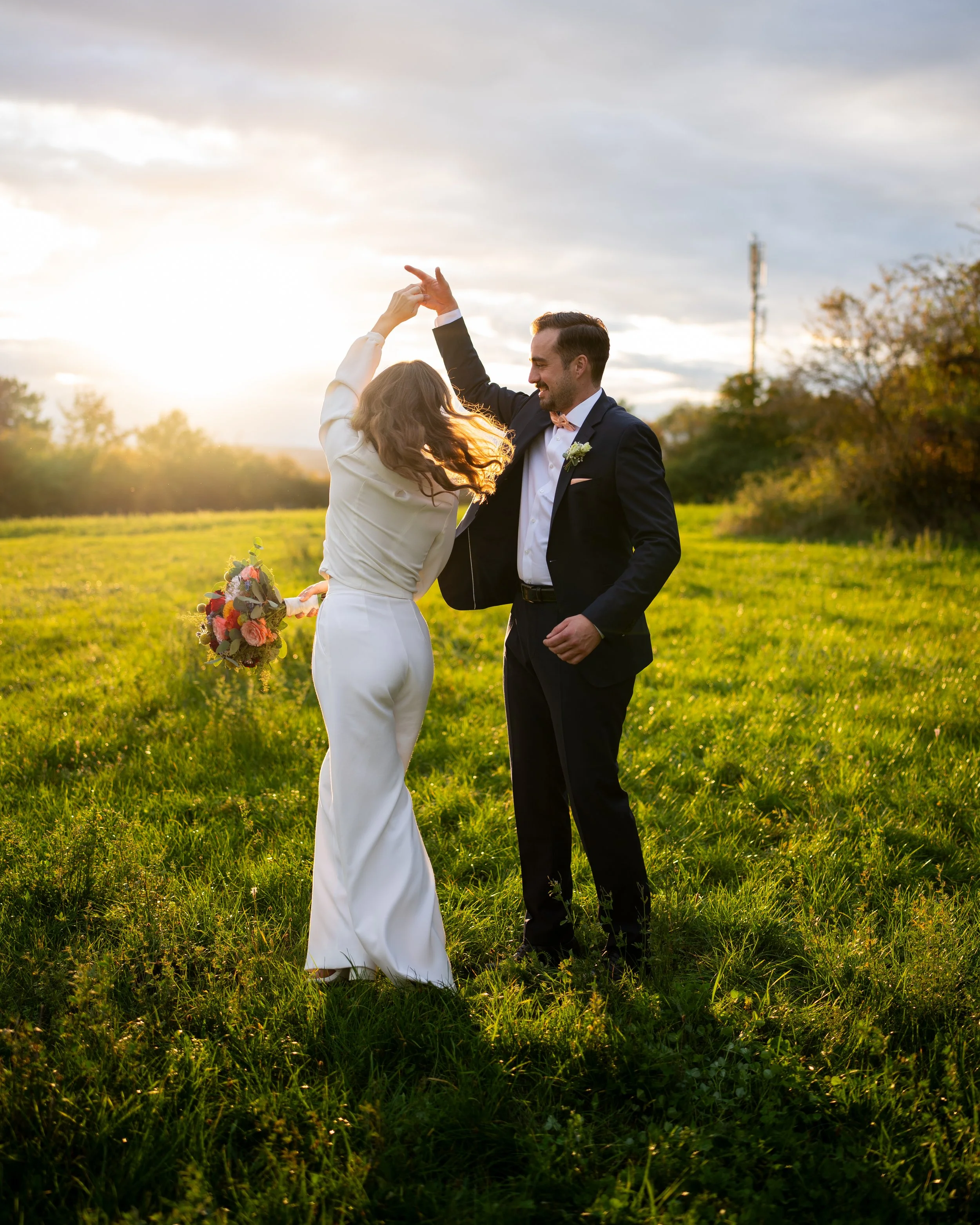 Ein Mann und eine Frau tanzen im Freien bei Sonnenuntergang, umgeben von grünem Gras und Bäumen, eine romantische Szene während einer Hochzeit.