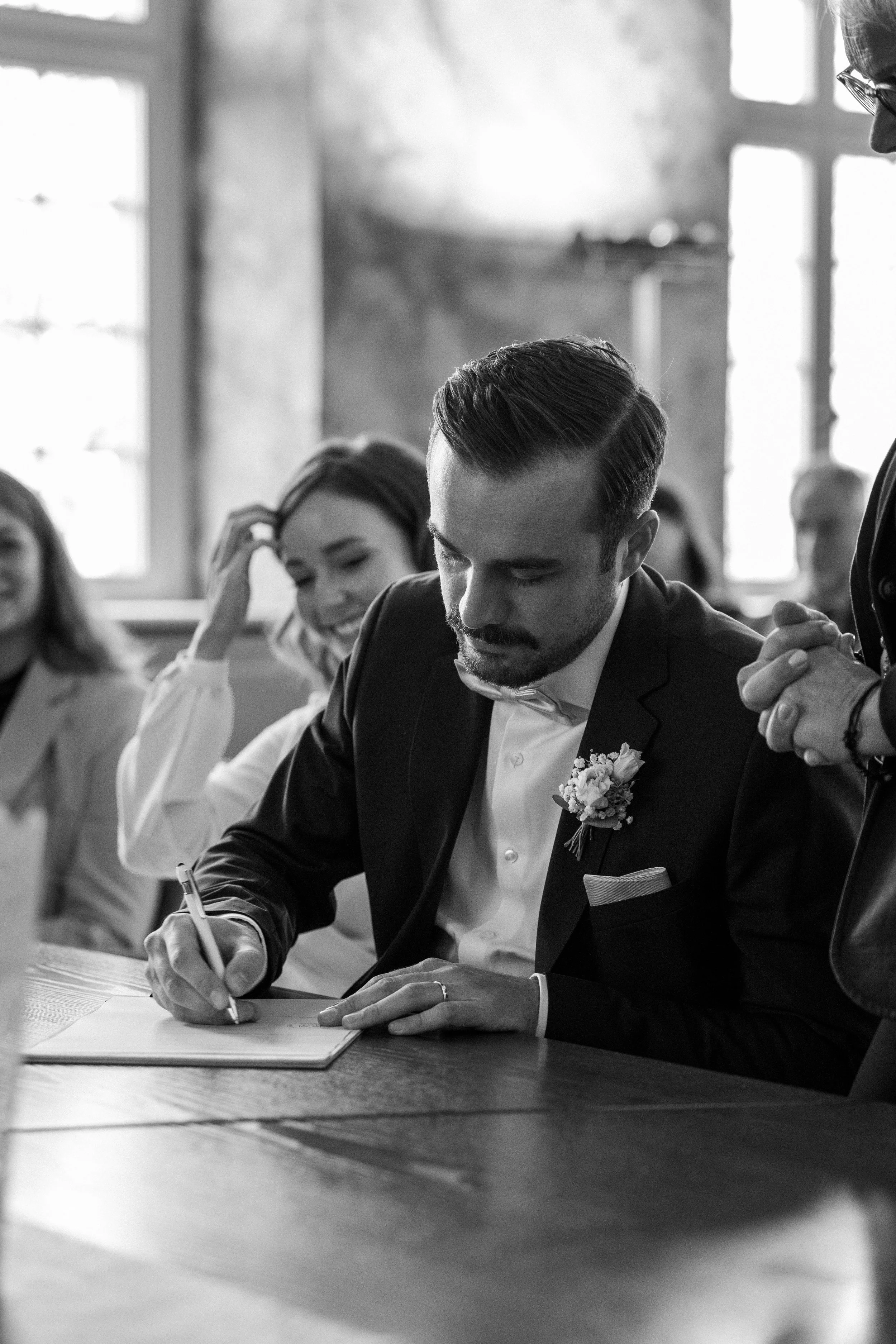 Ein Mann in einem schwarzen Anzug und weißem Hemd, der auf einem Papier mit einem Stift schreibt, während er in einem Raum mit großen Fenstern sitzt. Frauen sind im Hintergrund sichtbar, einige lachen.