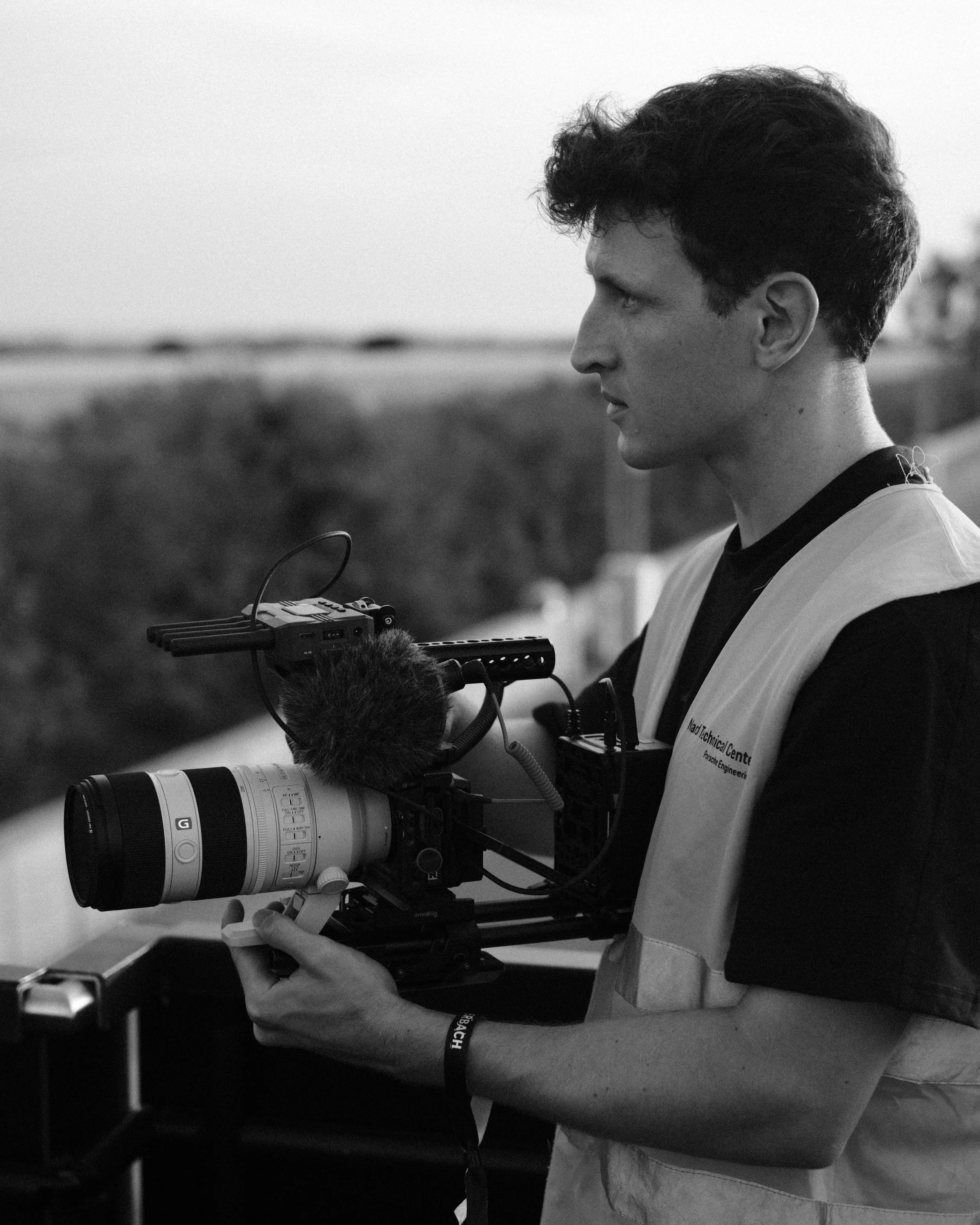 Ein junger Mann hält eine professionelle Kamera im Einsatz, auf einem outdoor Gelände bei Dämmerlicht, in schwarz-weiß Fotografie.