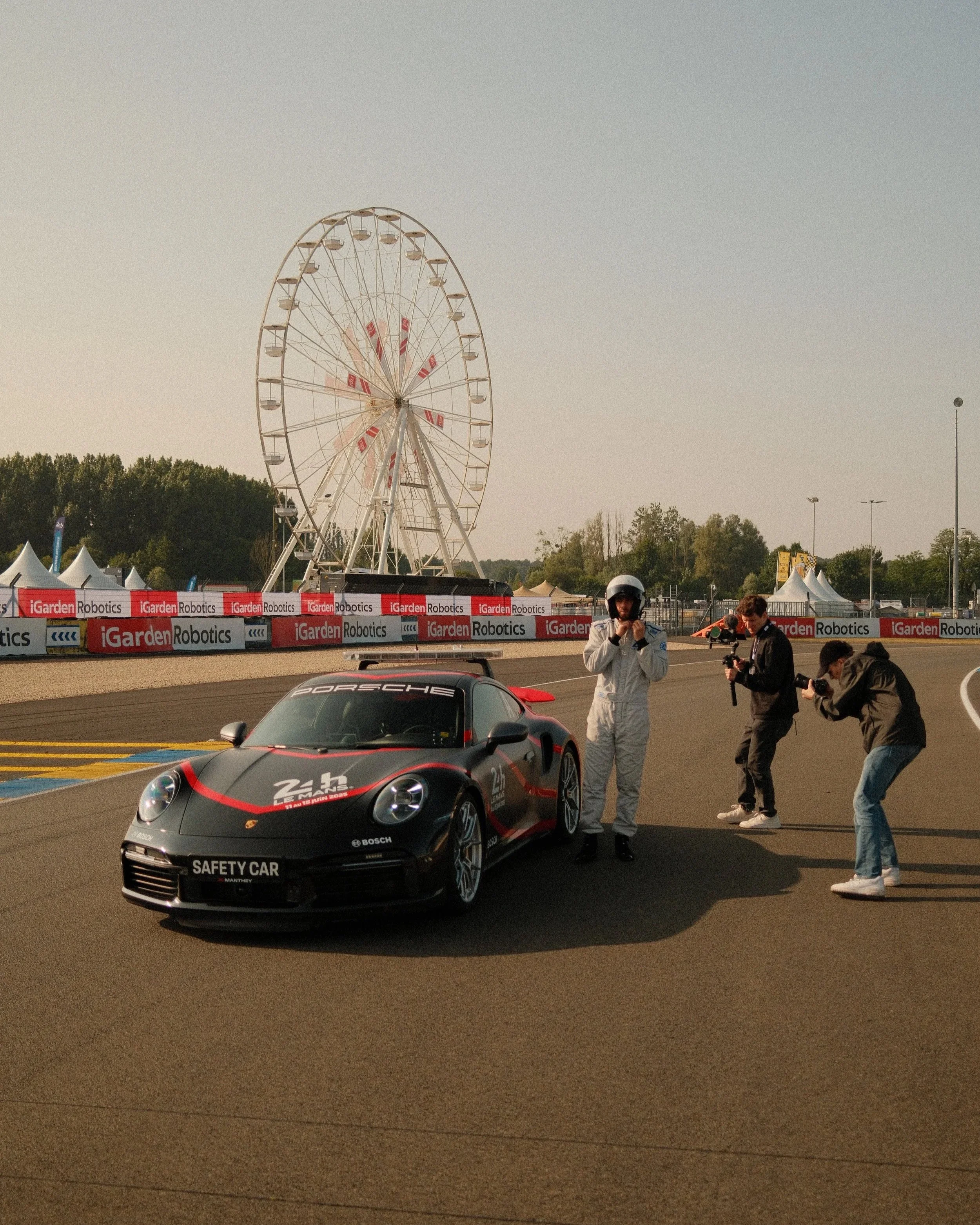 Ein schwarzer Porsche-Sportwagen mit der Aufschrift 'Safety Car' befindet sich auf einer Rennstrecke, im Hintergrund sind Menschen mit Helmen, Gebäude, ein Riesenrad, Zelte und Bäume sichtbar.
