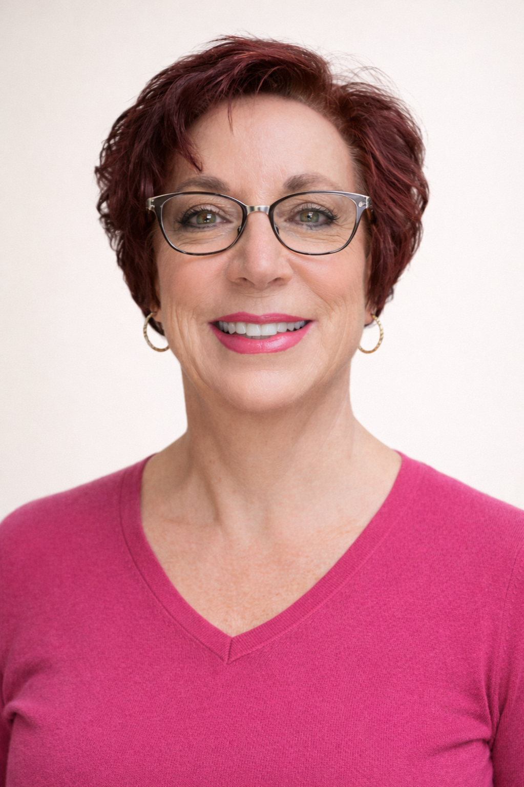 Close-up of a smiling woman with short red hair, glasses, earrings, wearing a pink top, against a light background.