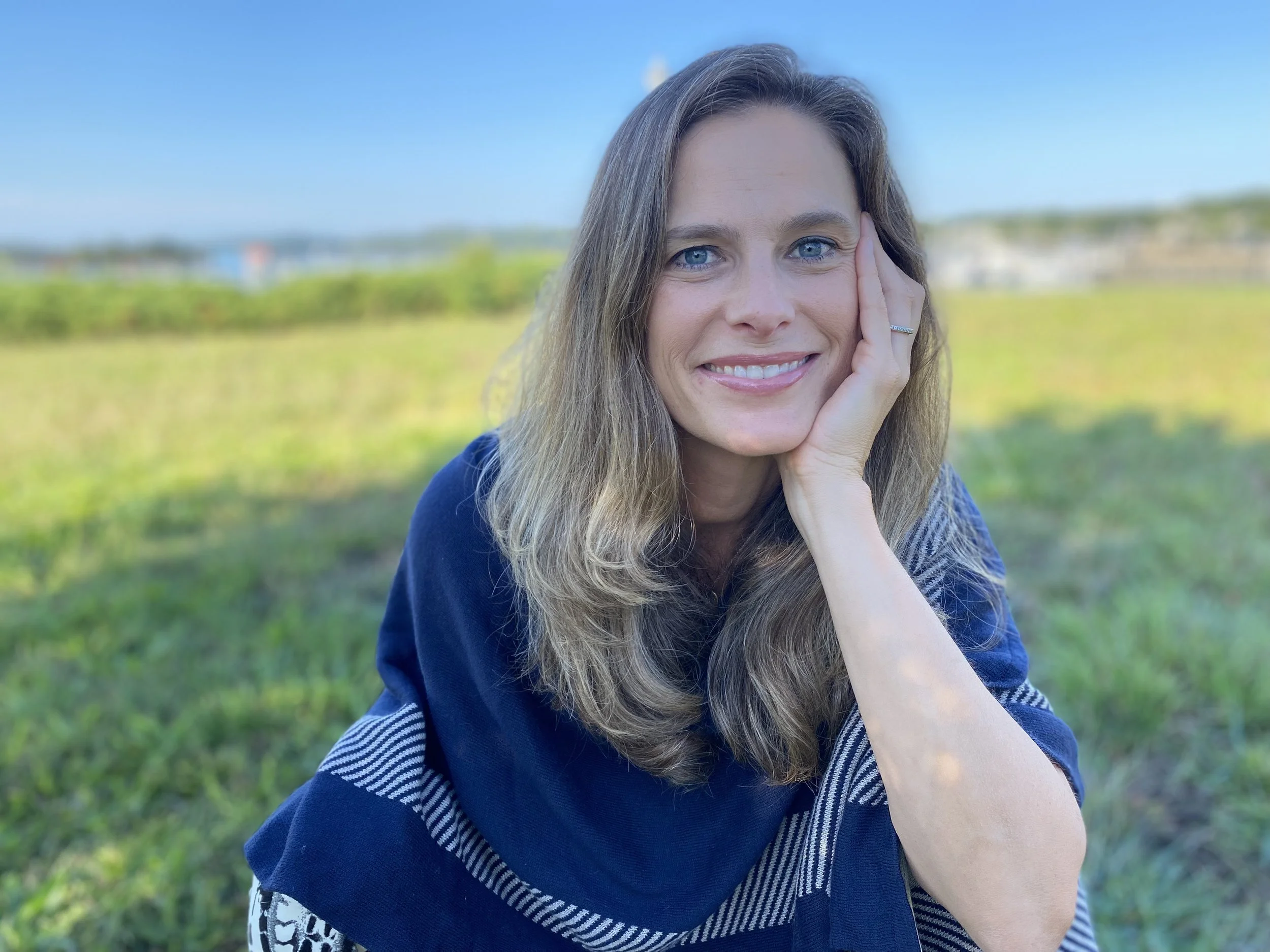 A woman with long, light brown hair and blue eyes smiling outdoors on a sunny day, resting her face on her left hand, with a blurred green landscape and blue sky in the background.
