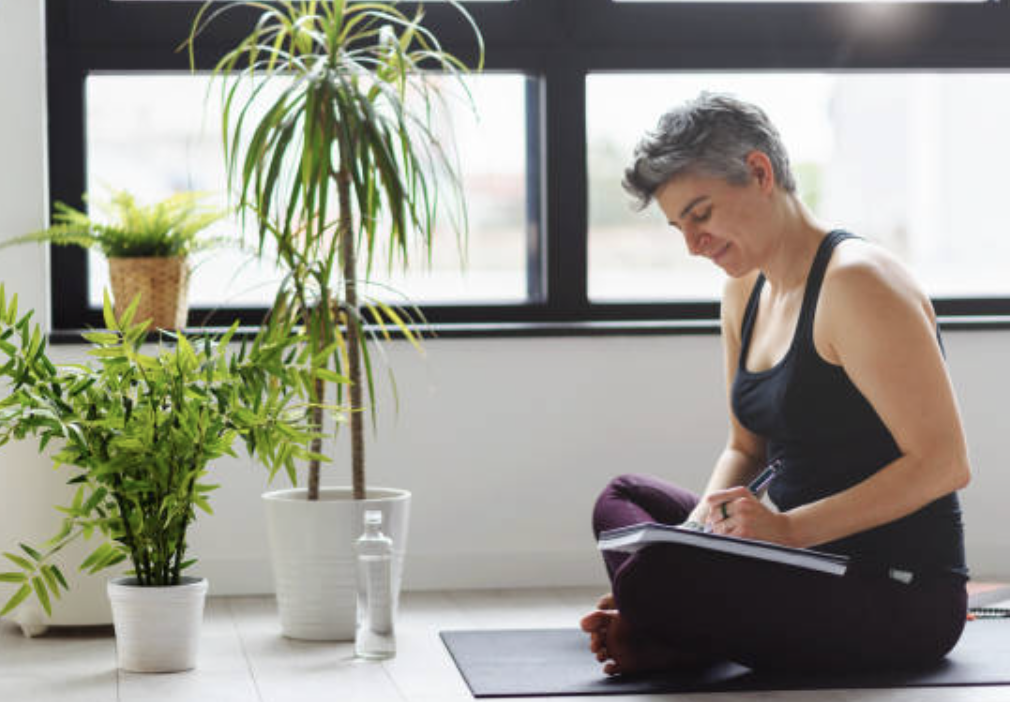 A woman sitting cross-legged on a yoga mat, writing in a notebook, with large green potted plants nearby and a window in the background.