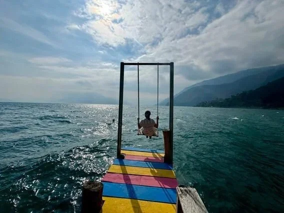 Person sitting on a swing on a colorful pier over a lake or ocean with mountains and cloudy sky in the background.