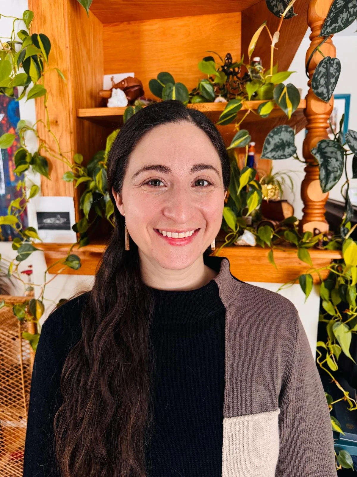 A woman with long dark hair, wearing a black, beige, and brown color-block sweater, smiling, standing in front of a wooden shelf with green plants and decorative items on it.