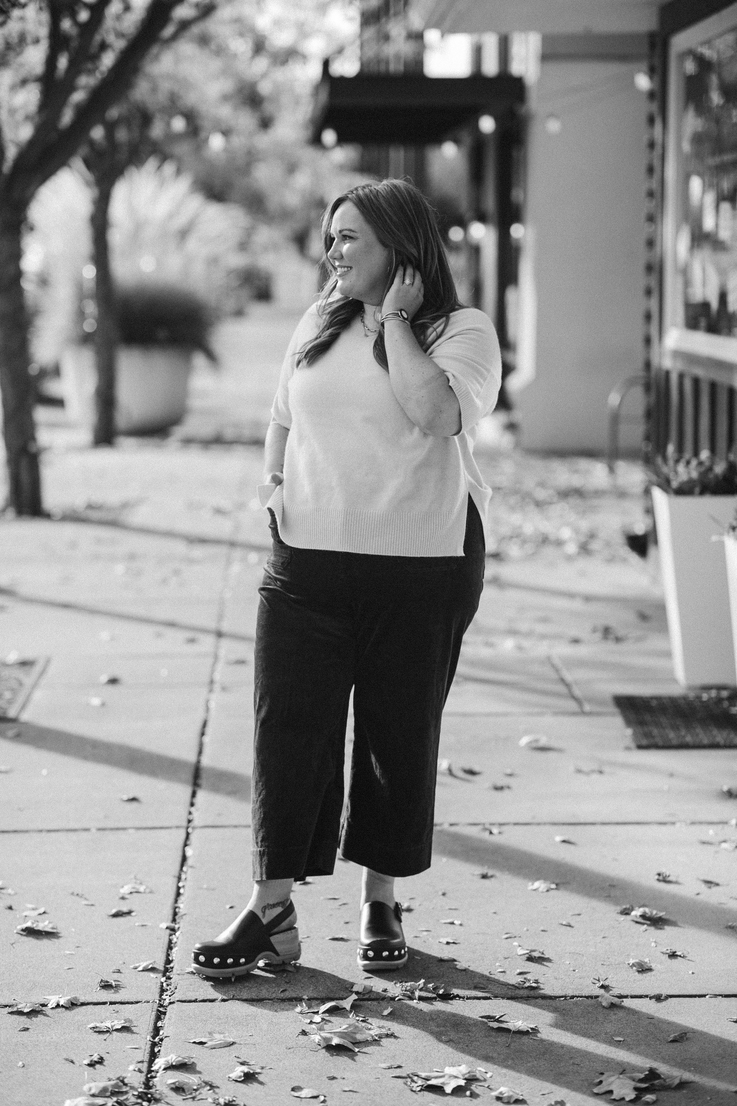 A woman with long hair, smiling and touching her ear, standing on a sidewalk surrounded by fallen leaves. She wears a light-colored sweater, dark wide-legged pants, and platform sandals. The background includes trees and storefronts, indicating an outdoor urban setting on a calm, autumn day.
