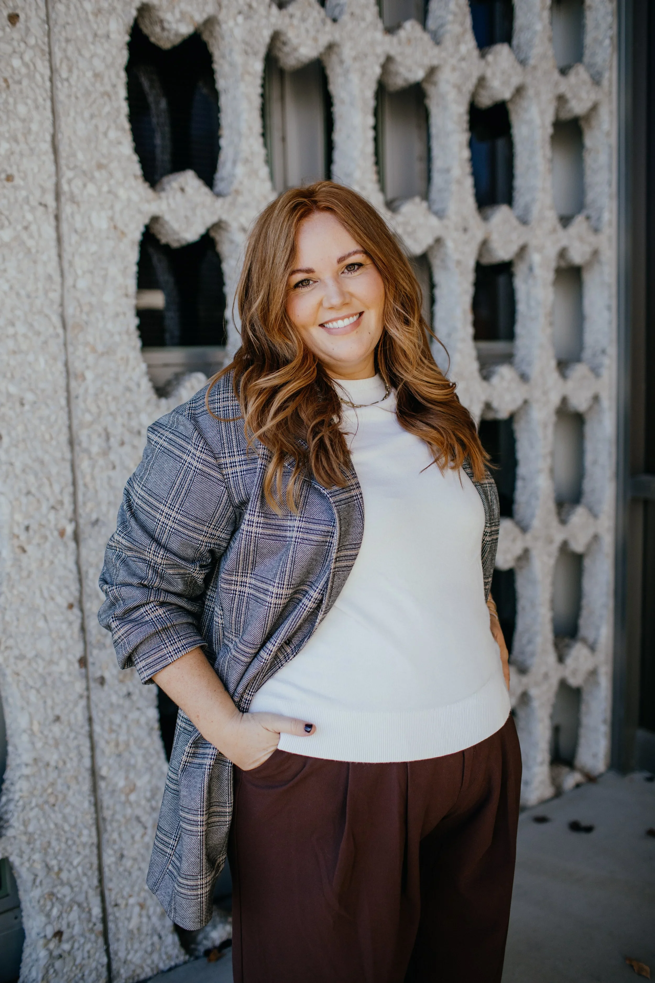 A woman with wavy brown hair, smiling, standing against a textured wall with geometric cutouts.