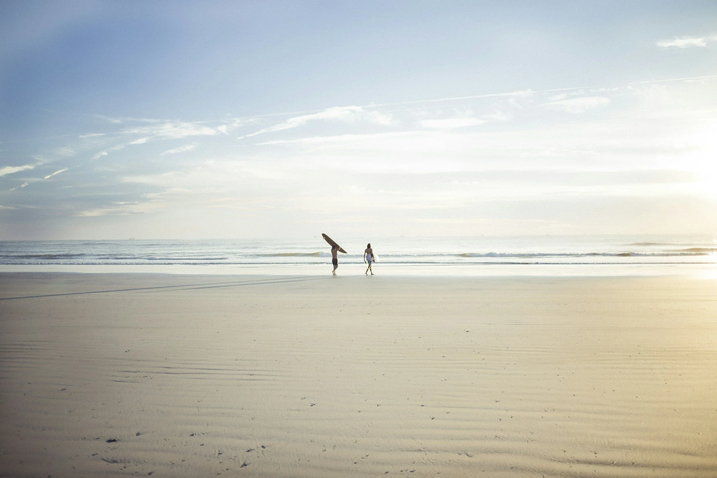 Two surfers walking on the beach with surfboards, under a bright sky with scattered clouds, gentle waves in the ocean.