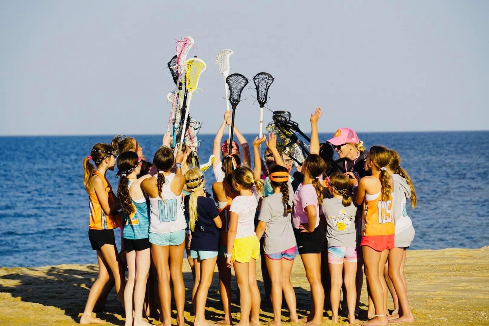 Group of girls in athletic wear with lacrosse sticks on a beach, participating in a team huddle or cheer near the ocean.