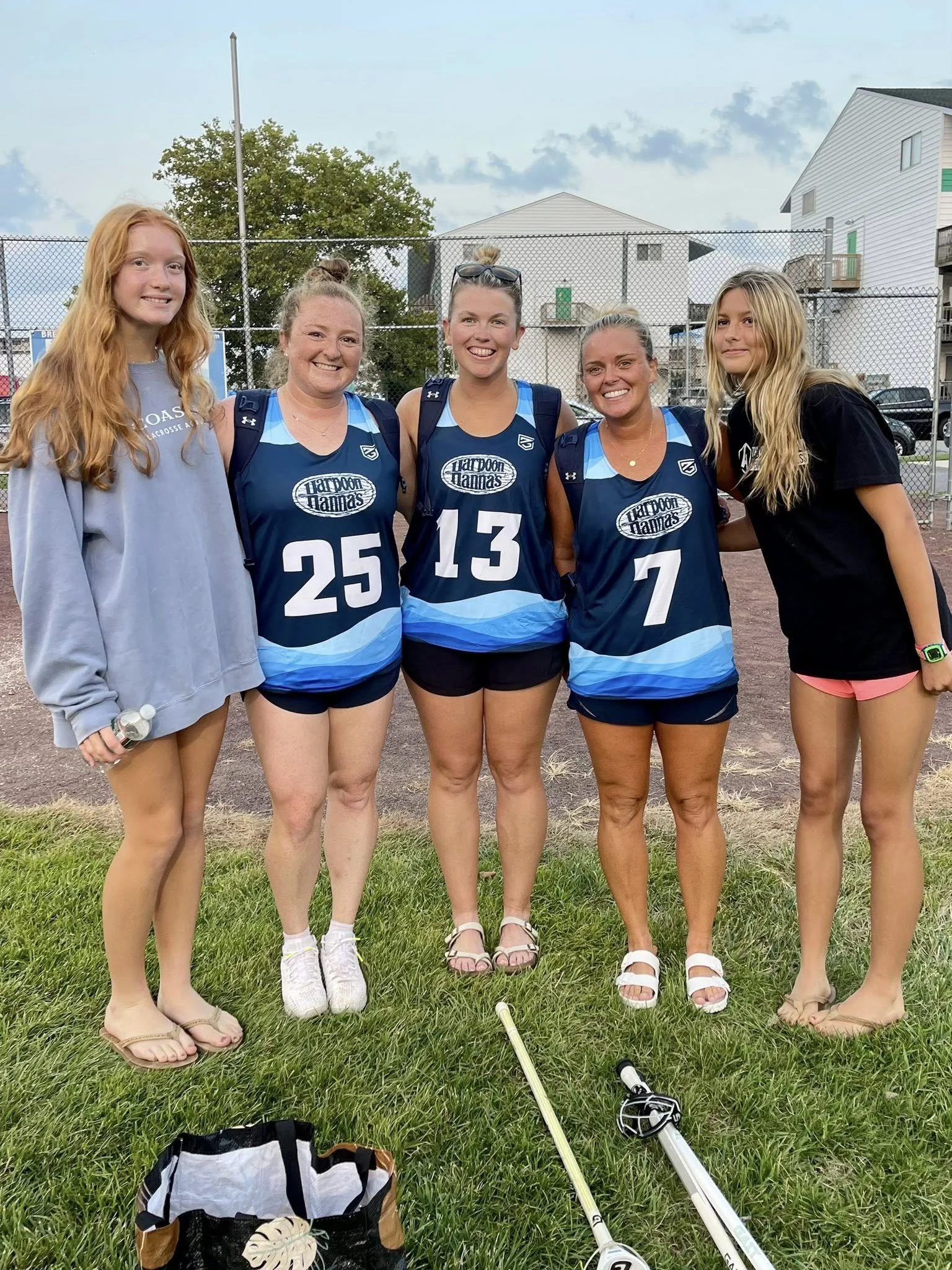 Five women standing outdoors on grass, smiling at the camera. Four are wearing blue sports jerseys with numbers and the words 'Haddon Hannan's' on them, and one woman on the far left is wearing a gray sweatshirt. There are beeping golf clubs and a bag on the ground in front of them, and a chain-link fence and residential buildings in the background.