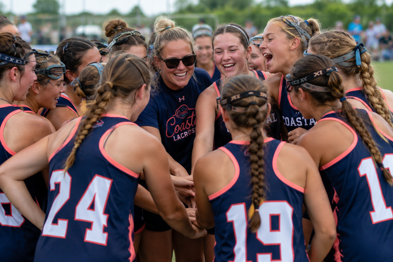 A group of female lacrosse players in navy blue and pink uniforms huddle together, smiling and laughing, with a coach or team leader in the center wearing sunglasses.