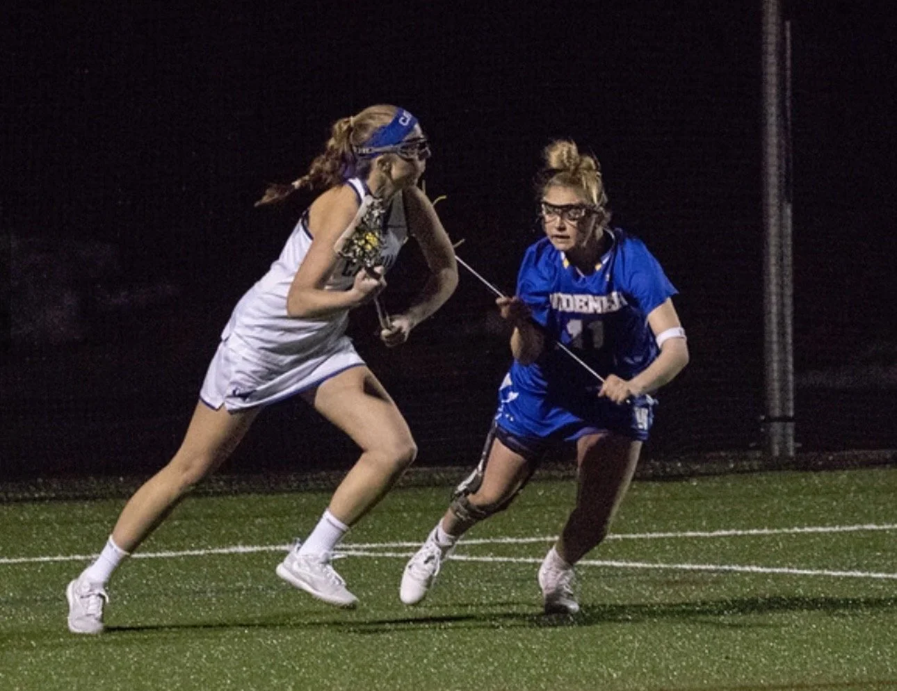 Two female field hockey players sprint on a dark, wet field at night, competing fiercely for control of the ball.